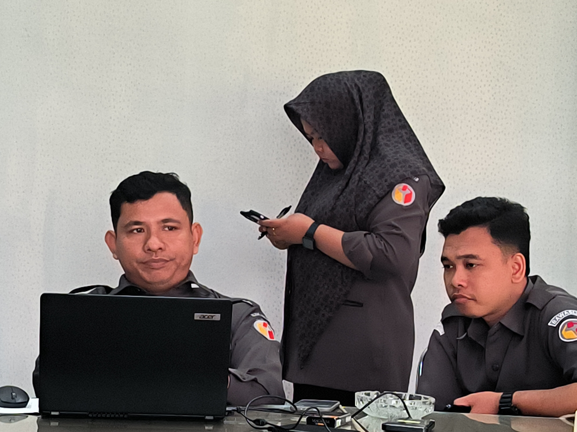 Three individuals are present in an indoor setting, possibly an office. A man seated on the left is positioned behind a laptop, with a computer mouse visible to his left. A woman stands in the center wearing a dark hijab. She appears to be holding and looking at a cellular phone. Another man is seated on the right. All three are wearing collared shirts with a circular patch with an emblem on the left breast. The location is purportedly in Lhokseumawe, Indonesia. Various items such as glasses, cables, and a small glass bowl are scattered on the surface in front of them. The background is a plain wall.