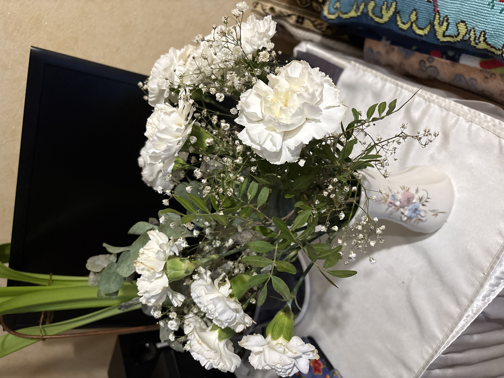 A close-up, indoor shot shows a bouquet of white carnations and baby's breath in a decorative vase. The vase has a floral pattern with blue and pink accents and is placed on a white tablecloth. Behind the vase, a dark monitor is visible, with a portion of a tan wall and what appears to be a patterned throw or fabric to the right. The lighting suggests an evening setting. The image does not contain any discernible elements to determine a specific location in Ternopil, Ukraine.