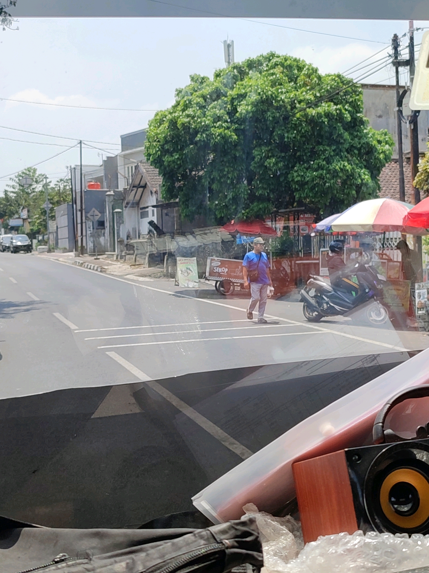 A street scene in Magelang, Indonesia, features a pedestrian crossing a road. The pedestrian, a man wearing a blue shirt, light-colored pants, and a cap, is walking from left to right across the road. He is carrying a bag over his shoulder and a white object in his right hand. A motorcycle is parked to the right of the pedestrian, and a food cart labeled "SEKOP" is visible behind him. Two colorful umbrellas, one red and white and another red, are set up near the food cart. The environment includes buildings, trees, and utility poles. The road is paved and has white lane markings. Reflections are visible on the surface from which the image was taken, possibly a vehicle window, obscuring parts of the scene. In the foreground, partially visible, are items like a black bag, a pink object, and a speaker.