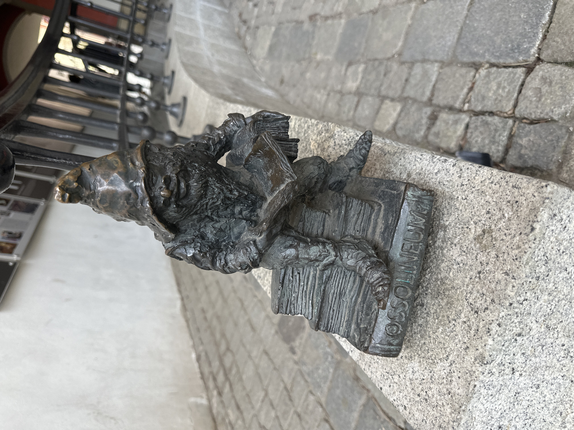 A bronze dwarf statue is affixed to the side of a stone wall in Wrocław, Poland. The dwarf is depicted wearing a pointed hat, with facial features of a wrinkled, gnome-like character. It's seated on a stack of books with the word "OSSOLINEUM" inscribed vertically. The dwarf holds a book in its hands and has a backpack or bundle slung over its shoulder. A metal railing with decorative spindles runs along the wall behind the statue. The wall is made of light gray textured stone, and the pavement below it is made of cobblestones.