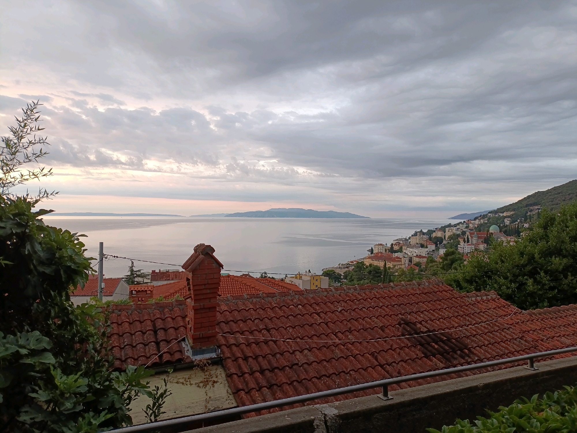 The image captures a panoramic view of the city of Opatija, Croatia, from an elevated vantage point. In the foreground, a weathered terracotta-tiled roof dominates, with a brick chimney standing prominently on its left side. Green foliage, including large-leafed plants, frames the left edge of the composition. Beyond the rooftops, the Adriatic Sea stretches out, its surface calm and reflecting the overcast sky. In the distance, a long, mountainous island is visible on the horizon. The cityscape of Opatija, with its colorful buildings and a distinctive green-domed structure, is nestled along the coastline, backed by lush green hills. The sky is filled with dramatic, gray clouds, suggesting either an impending storm or the aftermath of one, with a hint of soft light breaking through on the left horizon. There are no people or animals visible in the frame. The scene conveys a sense of peaceful, albeit somewhat moody, coastal life.