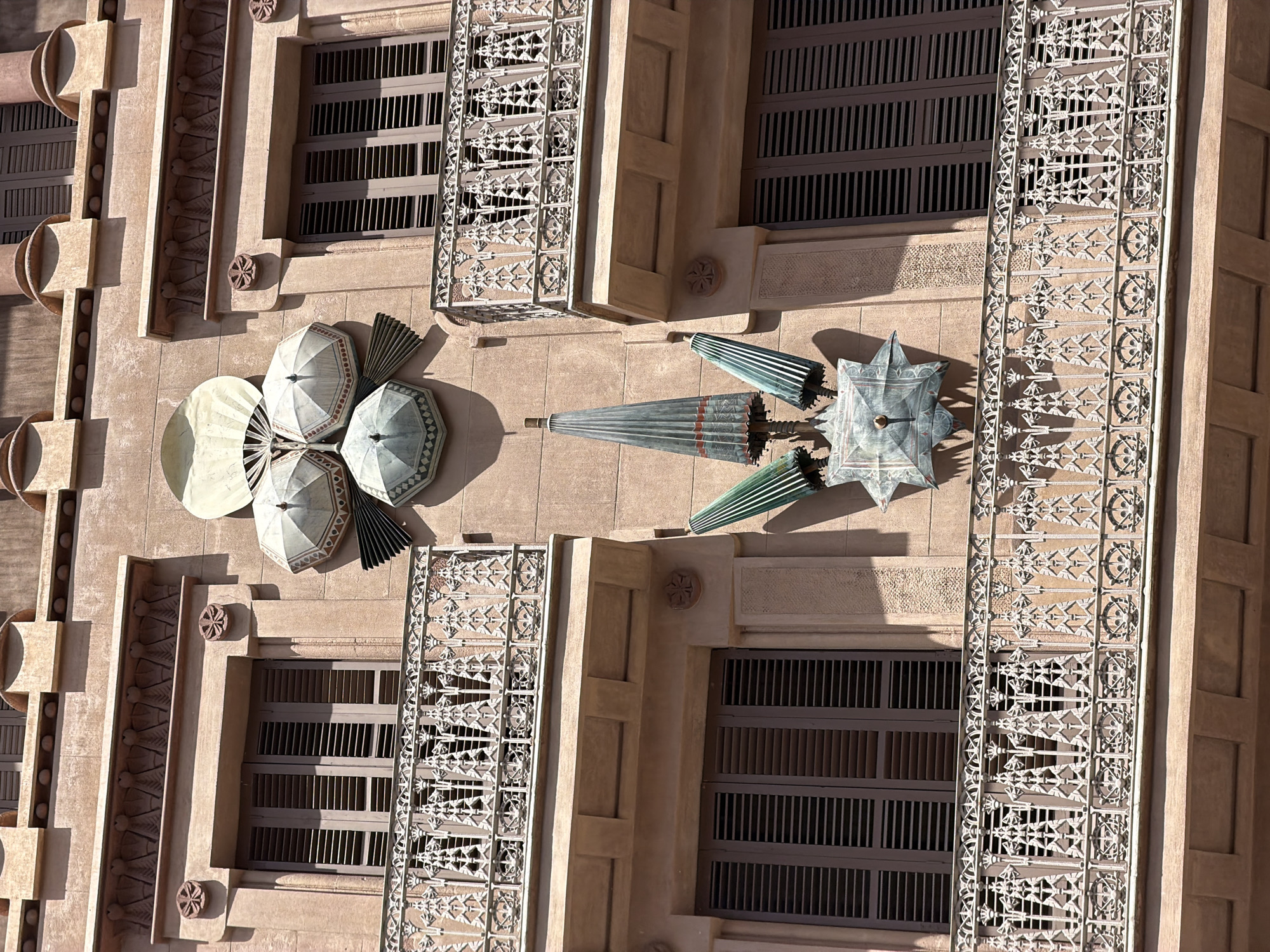 The image shows a close-up, eye-level view of the facade of a building in Barcelona, Spain, featuring decorative elements resembling umbrellas and fans. The building's exterior is a warm, sandy color, with windows featuring dark wooden shutters. Intricate wrought-iron railings adorn balconies that extend from the building.
The central decorative feature is a cluster of metallic objects shaped like opened and closed umbrellas, along with what appear to be decorative fans. Some of these objects are arranged in a radial pattern, with a larger, fan-like element at the top and several umbrella-like structures below, some partially open and others closed. Further down, another arrangement of closed, elongated umbrella shapes points downwards, attached to a star-shaped metallic piece. The metallic elements have a greenish patina, suggesting they are made of copper or brass that has oxidized.
The architectural style appears to be a blend of Art Nouveau and possibly other ornamental styles, with decorative friezes above the windows and circular rosettes on the pilasters. The sunlight casts shadows on the building, highlighting the textures and three-dimensional aspects of the ornamentation. There are no people visible in the image. The building is likely residential or commercial, judging by the windows and balconies. The specific location is not provided in the image itself, but similar decorative elements are characteristic of buildings designed by architects like Antoni Gaudí in Barcelona.