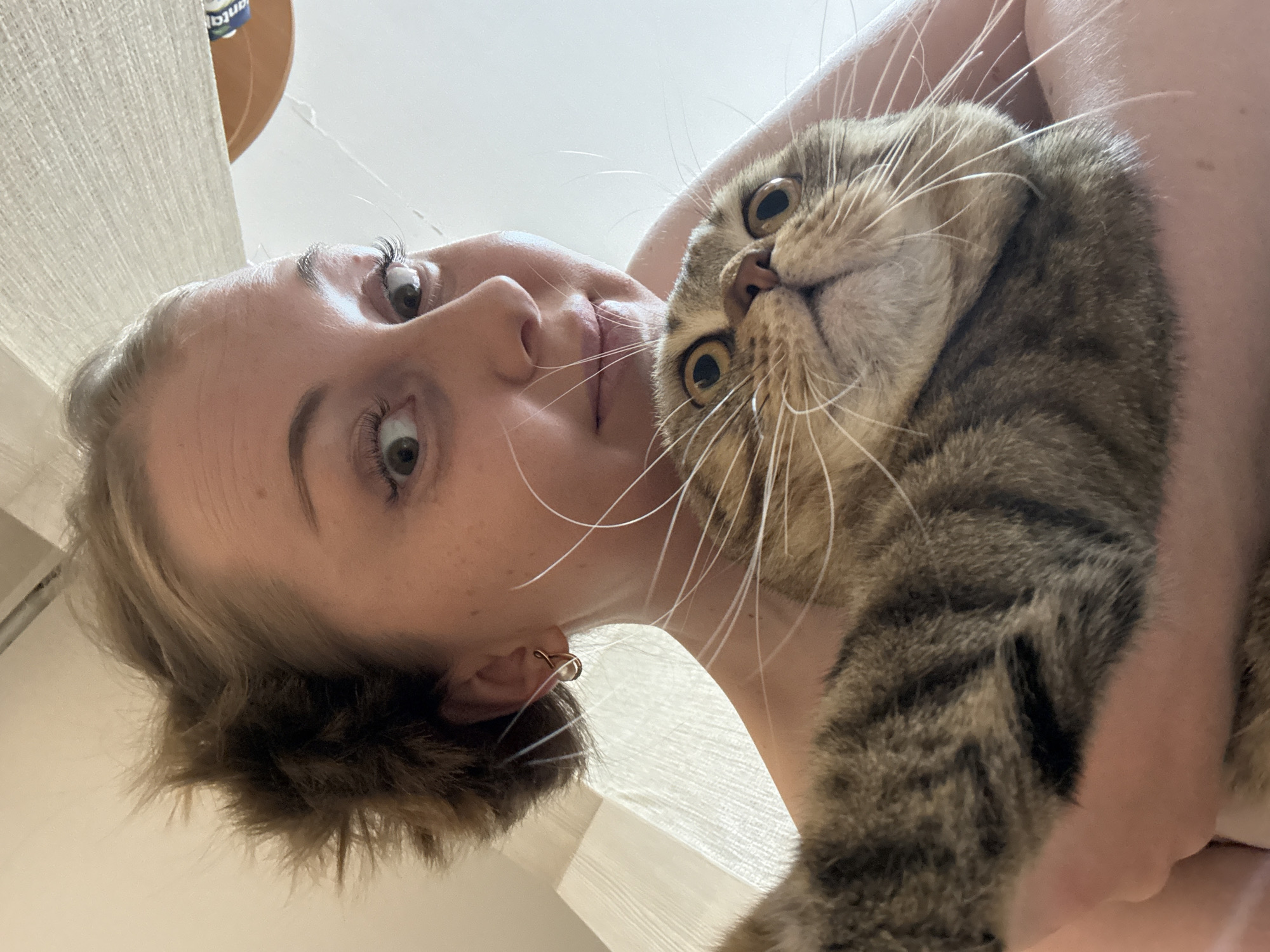 A young woman with blonde hair and an earring in her left ear is holding a tabby cat. The cat is looking up at the camera with wide eyes. The background is white. The location is unknown.