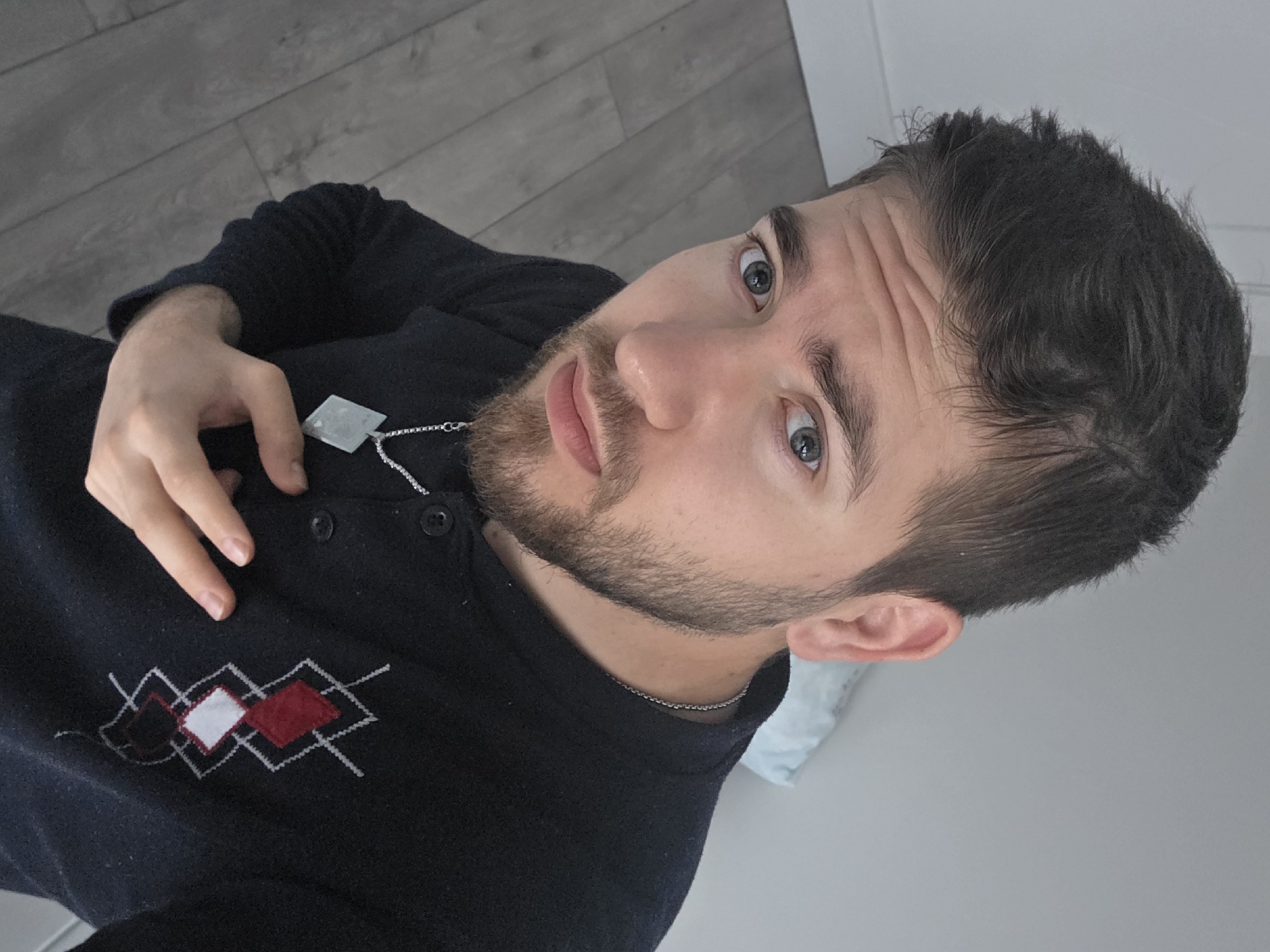 A fair-skinned man with short dark hair, a beard, and light-colored eyes is positioned at an upward angle. He is wearing a dark-colored top featuring an argyle pattern on the left side of his chest and a silver pendant. The background includes gray wood flooring and a light-colored wall. The location, Yagodovo, Bulgaria, cannot be determined from the visual information.