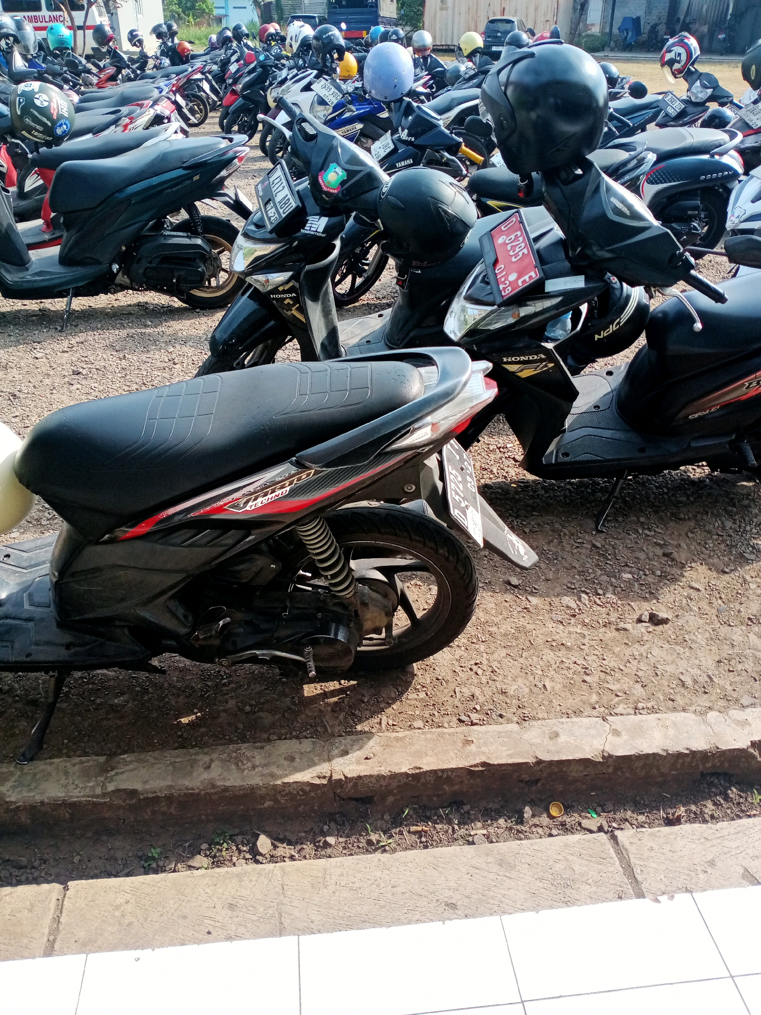 The image shows a parking lot filled with many scooters, primarily Honda brand, parked on a gravel surface. The scooters are predominantly black and various shades of gray, with some accent colors like red and yellow. Many of the scooters have helmets placed on them or nearby. In the background, a white ambulance is visible, suggesting the location might be near a hospital or clinic. The overall scene depicts a typical urban or suburban parking area in Indonesia, possibly in Cangkuang Wetan, given the contextual information. The environment is outdoors, with sunlight casting shadows.