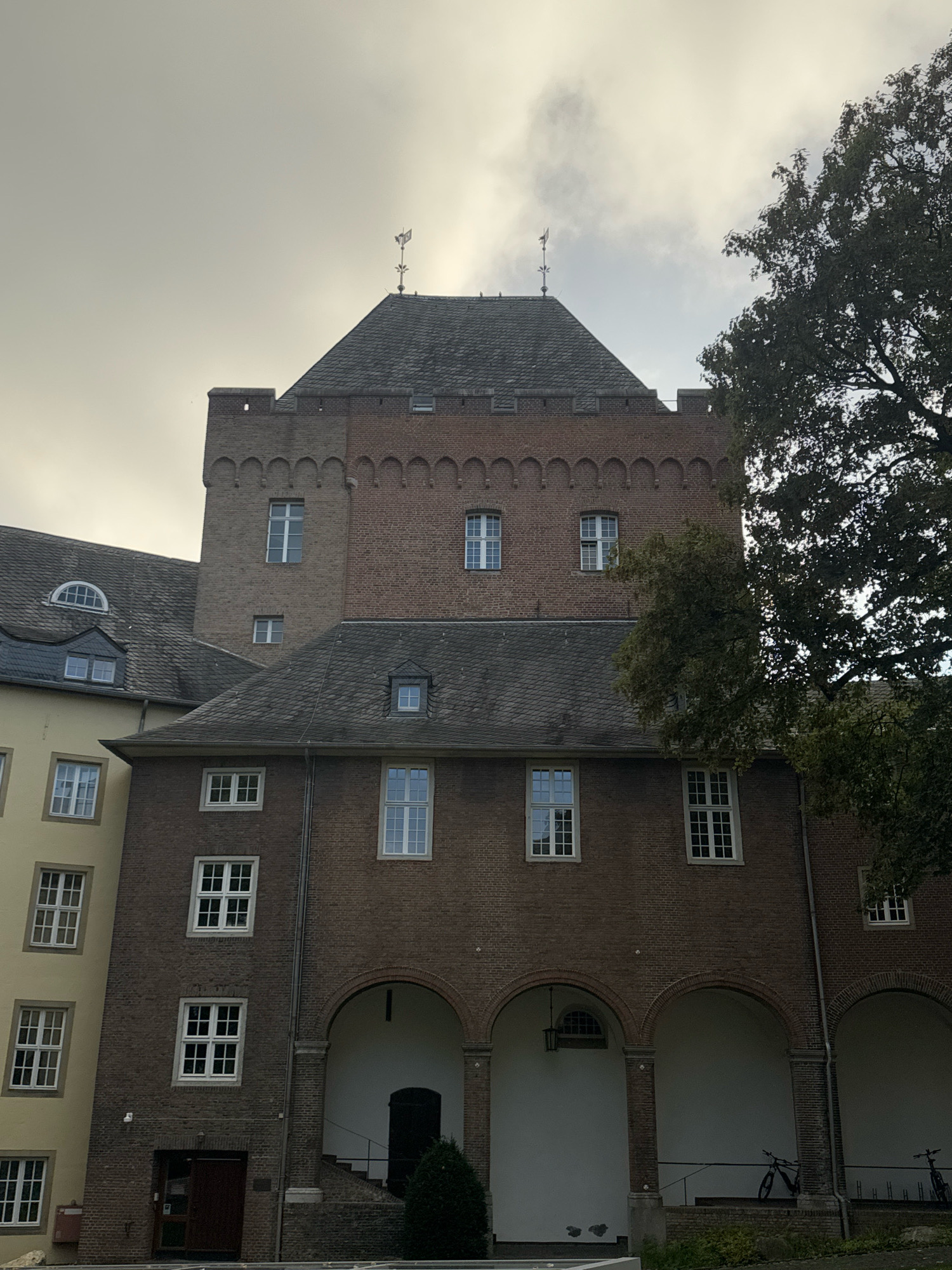 The image shows the exterior of a brick building in Cleves, Germany. The building has multiple stories with a dark, tiled roof. There are numerous windows of various sizes and styles visible. A tall, darker brick tower is integrated into the main structure, featuring decorative brickwork at the top and crenellations. A portion of a large, dark green tree is on the right side of the image, partially obscuring the building. The sky above is overcast with some lighter clouds. In the foreground, an arched walkway with columns is visible, and two bicycles are parked within the arched area. There are no people visible in the image. The building appears to be an older, possibly historic structure.