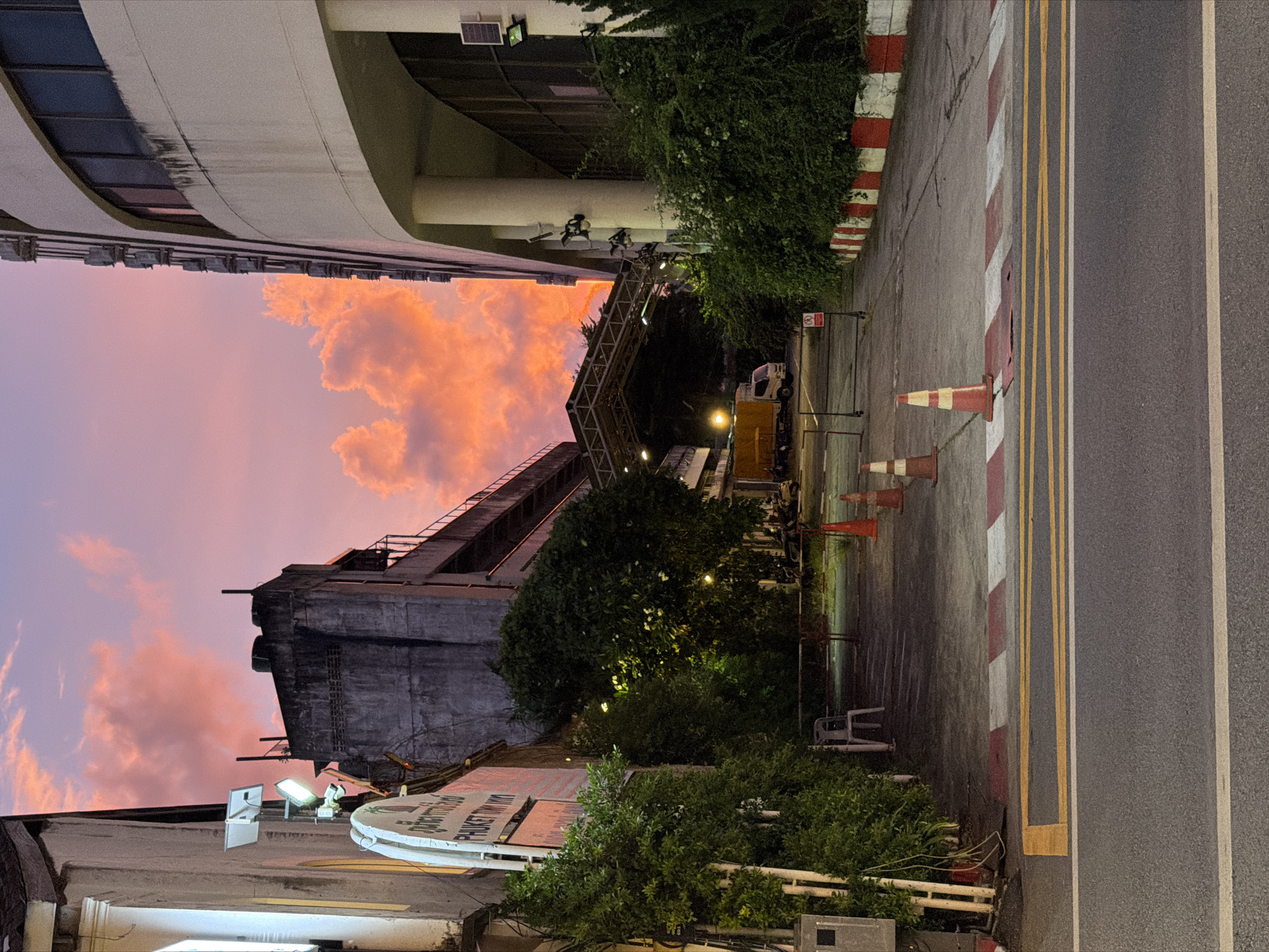 The image depicts a street scene at dusk in Phuket City Municipality, Thailand. The sky is dominated by a vibrant sunset, with dramatic pink and orange clouds illuminated against a fading purple sky. In the foreground, a paved road with yellow and white lane markings is visible. A raised crosswalk with red and white striped barriers is also present. The scene is flanked by buildings on either side. On the left, a multi-story building with a white facade and signage is partially obscured by dense green foliage. On the right, a large, curved, light-colored building with multiple windows and exterior air conditioning units stands tall. The middle ground shows an alleyway or driveway leading between the buildings, with more greenery and a covered structure. A yellow truck is parked in the distance. Several orange traffic cones are scattered along the side of the road and in the driveway. A white plastic chair is placed near the left-hand building, next to a metal barrier. The lighting suggests it is either early evening or late twilight, with artificial lights from the buildings and street lamps beginning to illuminate the scene.