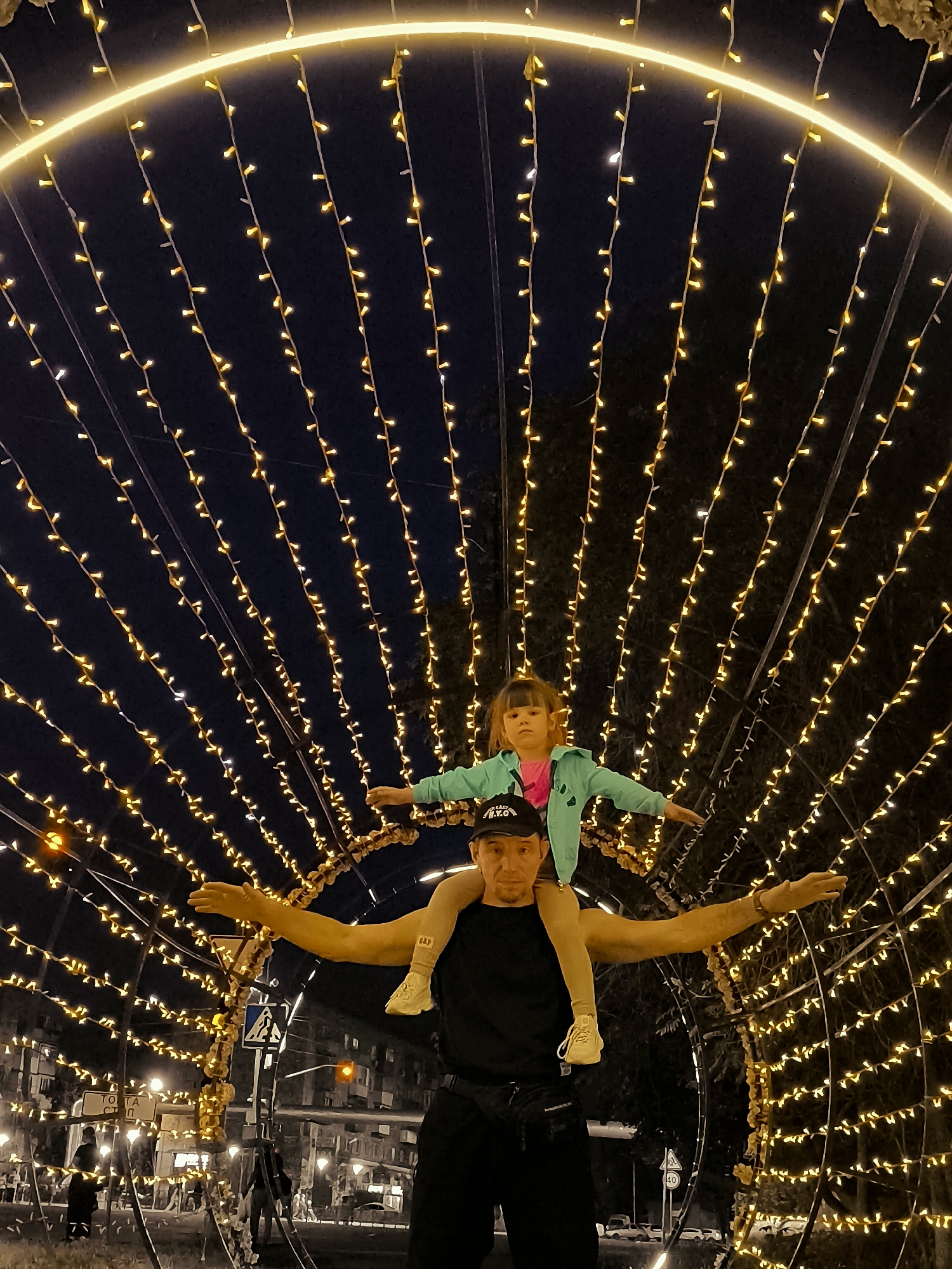 A man carries a young child on his shoulders in front of a large archway decorated with strings of yellow lights. The man, wearing a black t-shirt and a black baseball cap, faces the camera with a neutral expression. The child, wearing a turquoise jacket over a pink shirt, has their arms outstretched and is looking to the side. The archway creates a tunnel-like effect, with the lights extending upwards and outwards in a symmetrical pattern against a dark night sky. In the background, blurred lights and the silhouettes of buildings suggest an urban setting. Traffic lights and street signs are also visible. The overall mood is festive and playful, likely taken at night during a holiday or special event. The text "N.Y.C." is visible on the man's baseball cap.