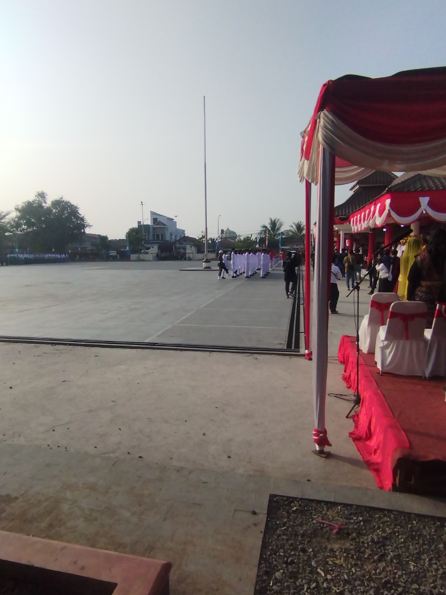 The image depicts an outdoor event, likely a ceremony or parade, taking place in Kramatwatu, Indonesia. The foreground shows a concrete area and a section of a decorated stage with red fabric draping and white chairs. In the midground, a large, flat, paved area, possibly a field or square, is visible. A line of individuals in white uniforms is marching or standing at attention on this paved area. To the left, trees and buildings can be seen in the background. The sky is overcast and bright. A tall flagpole stands prominently in the distance. Several people are observing the event from the area near the stage, some appearing to be participants or officials, including one individual in a yellow headscarf. The overall scene suggests an organized public gathering under a bright, possibly humid, tropical sky.