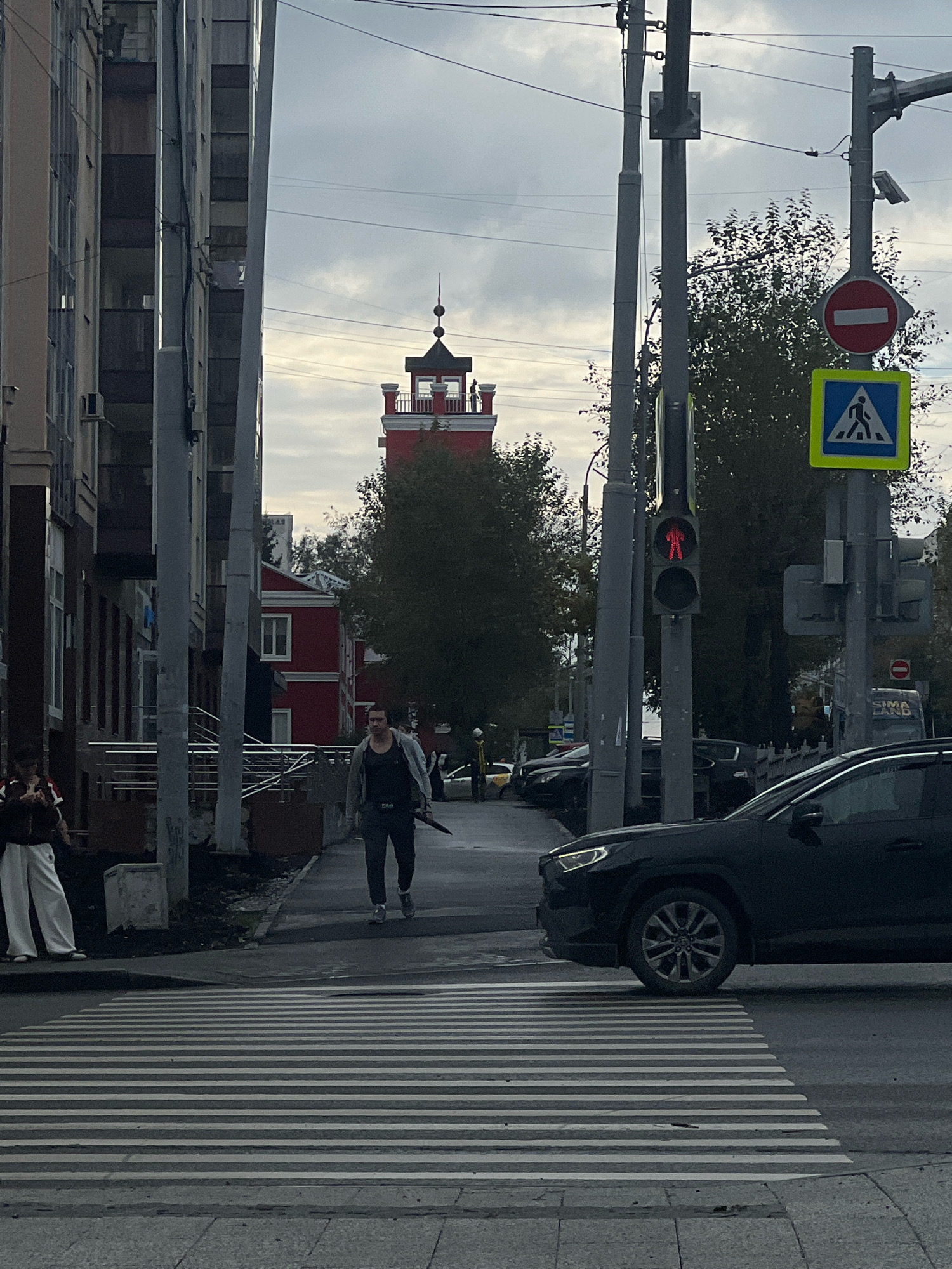 A man is walking across a pedestrian crossing on a cloudy day in Yekaterinburg, Russia. The man is wearing a dark tank top, a light-colored jacket, dark pants, and grey shoes. He is carrying a dark, umbrella-like object. He is walking from left to right in the frame, towards a red building with a dark tower on top. The pedestrian crossing is wide and marked with white stripes. To the right of the crossing, a black SUV is partially visible, with its front wheel on the crossing. In the background, there are tall buildings on the left, power lines crisscrossing the sky, and trees. Traffic lights are visible, with a red pedestrian signal illuminated. Several traffic signs are also present, including a "no entry" sign and a pedestrian crossing sign. The overall atmosphere is urban and somewhat overcast.