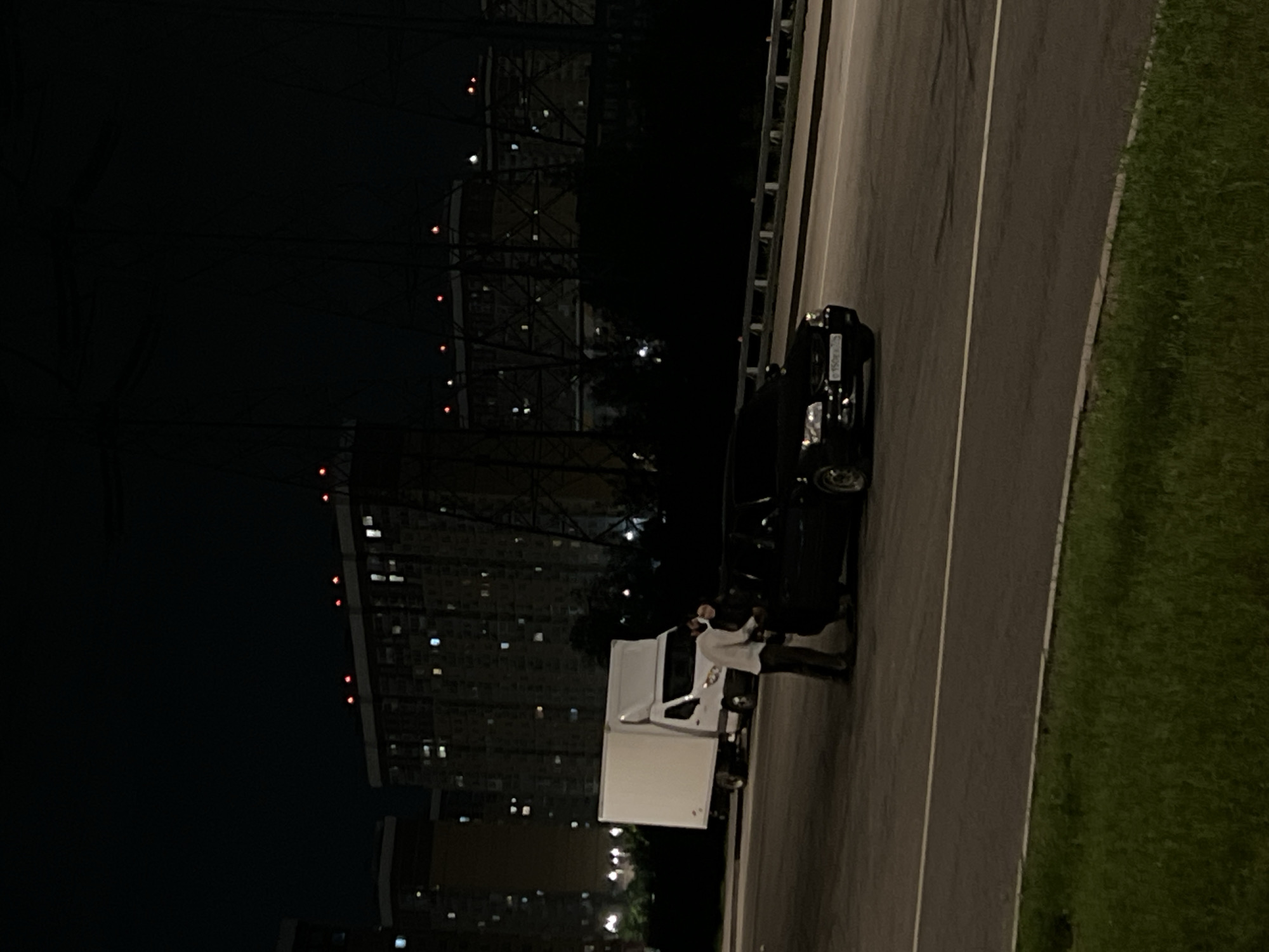 The scene depicts a street in Moscow, Russia at night. The illumination is dim, with artificial lights providing the primary light source. A white box truck is parked on the right side of the road next to a black vehicle. A person standing by the driver-side door of the box truck appears to be interacting with someone inside. The road runs parallel to a grassy area on the right. In the background, tall buildings with illuminated windows are visible. Additionally, structural towers, possibly part of a bridge or some other infrastructure element, are visible in the night sky. The overall environment seems urban with a mix of residential or commercial structures and infrastructure.