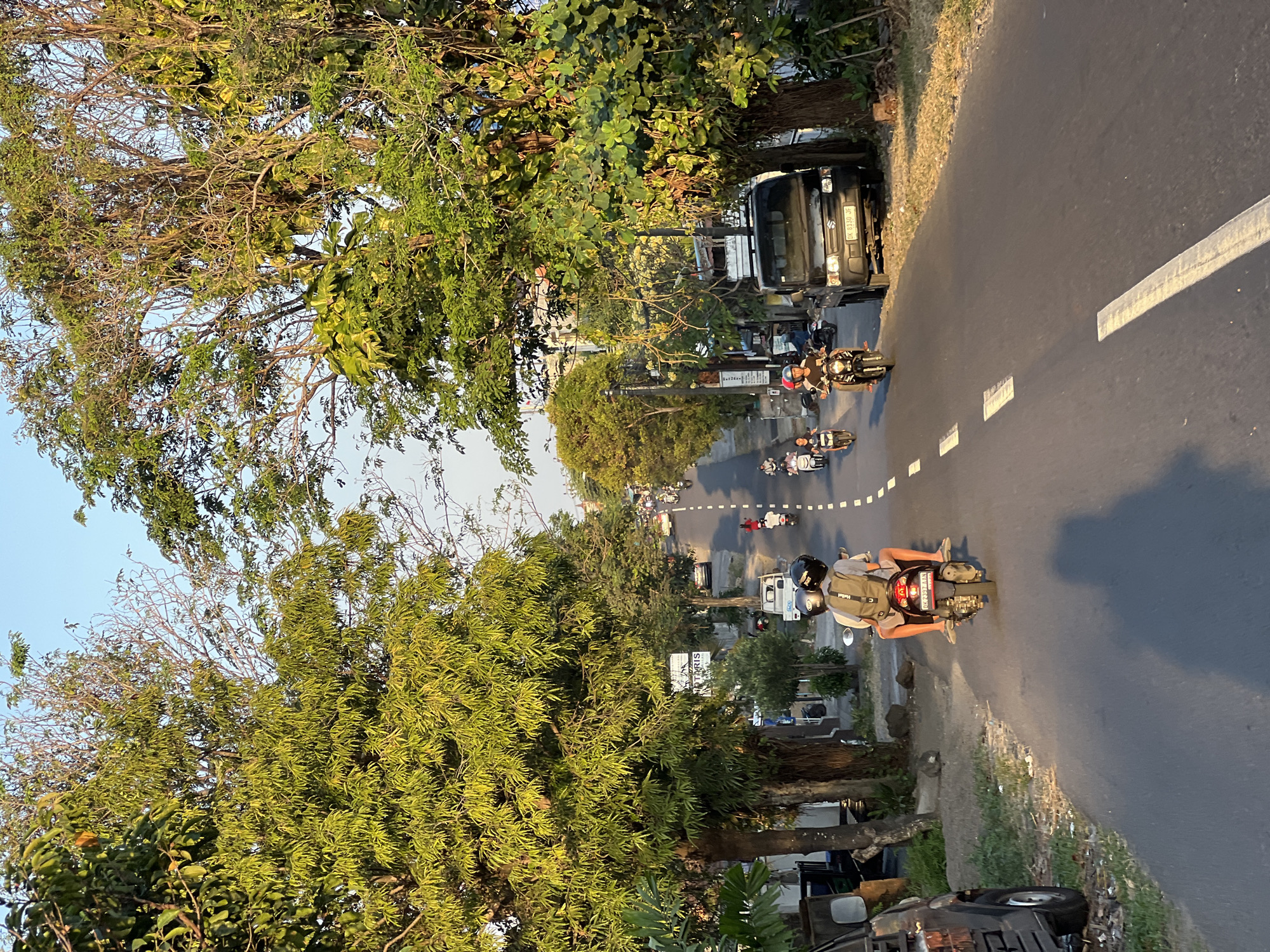 This is a scene captured in Denpasar, Indonesia, on a paved road lined with trees. The road slopes slightly upwards, with a white dashed line marking the center. Several motorcycles are in motion, with riders and passengers navigating the street. In the foreground, two individuals are riding a motorcycle, with the passenger wearing a backpack. A dark-colored pickup truck is parked on the right side of the road. Further up the road, more motorcycles and vehicles are visible, along with buildings and signs on the left side, including one that appears to be for a business. The sunlight casts long shadows across the road, indicating it is likely late afternoon or early evening. The overall environment suggests a typical urban or suburban street scene in Indonesia.