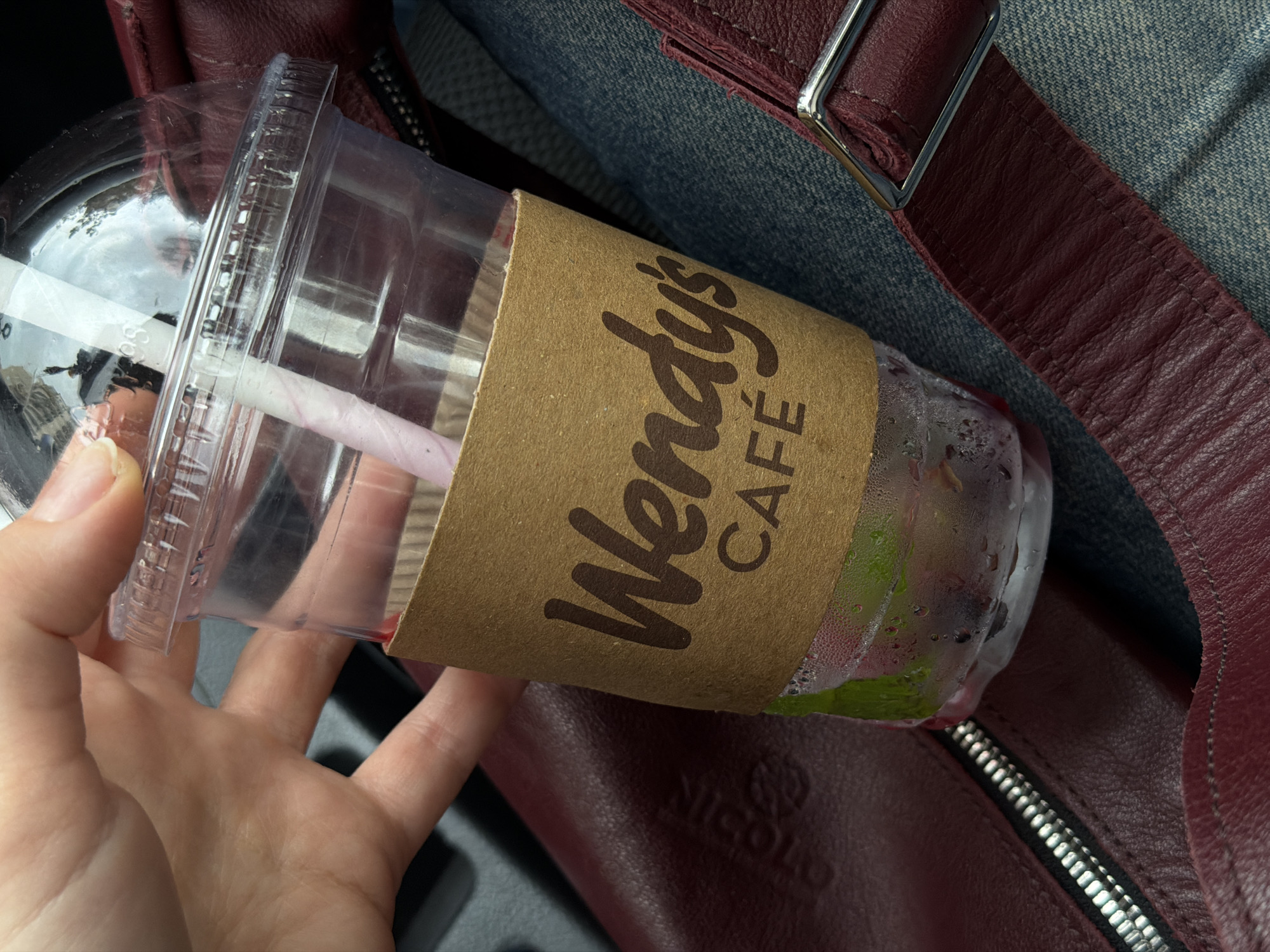 A hand holds a clear plastic Wendy's Cafe cup with a brown cardboard sleeve. Inside the cup, a pink and white striped straw is visible amidst a beverage that appears to contain ice and possibly fruit or other ingredients. The cup shows signs of condensation. The cup sits on a maroon leather bag with a silver zipper and a silver buckle on a strap, with a blue jean-like material in the background. The location is Tbilisi, Georgia, implied by a subtle, indistinct reflection in the plastic cup.