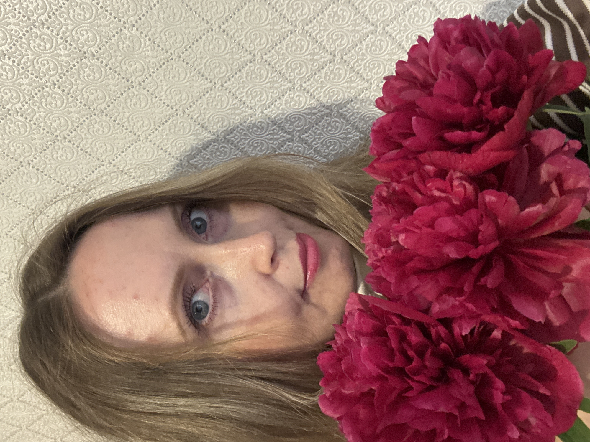 A close-up shot features a woman and three vibrant deep pink peonies. The woman's face is positioned on the left side of the frame, with her blonde hair cascading around her. She has fair skin, wide blue eyes, and is wearing light pink lipstick, with a slightly surprised or curious expression. A subtle sheen is visible on her forehead.
To the right, three large, fully bloomed peonies showcase a rich magenta or fuchsia color with numerous ruffled petals. The flowers appear to be held or placed close to the woman's face, creating a strong contrast with her fair complexion.
The background consists of a light-colored, possibly cream or white, wall with an ornate, embossed pattern of interlocking scrolls and diamond shapes filled with tiny dots, suggesting wallpaper or a decorative panel. Subtle shadows on the wall indicate indoor lighting. No visible text is present in the image. The scene is located in Chaikovsky, Russia.