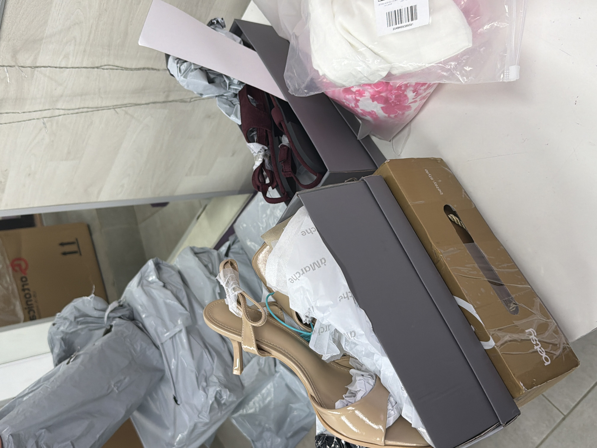 The image presents a collection of items placed on a white surface, likely a table or counter. There are several shoe boxes, one labeled "betsy", alongside a pair of light beige heeled sandals and a pair of burgundy shoes. The shoes appear to be partially unpacked. A cardboard box with "orouics" printed on it is also visible. The scene includes white tissue paper and plastic packaging, suggesting recently opened or new items. The location is indoors, with what appears to be a reflection in a mirror in the lower left corner of the image. The city of Moscow, Russia, is not discernible from the image's visual information.