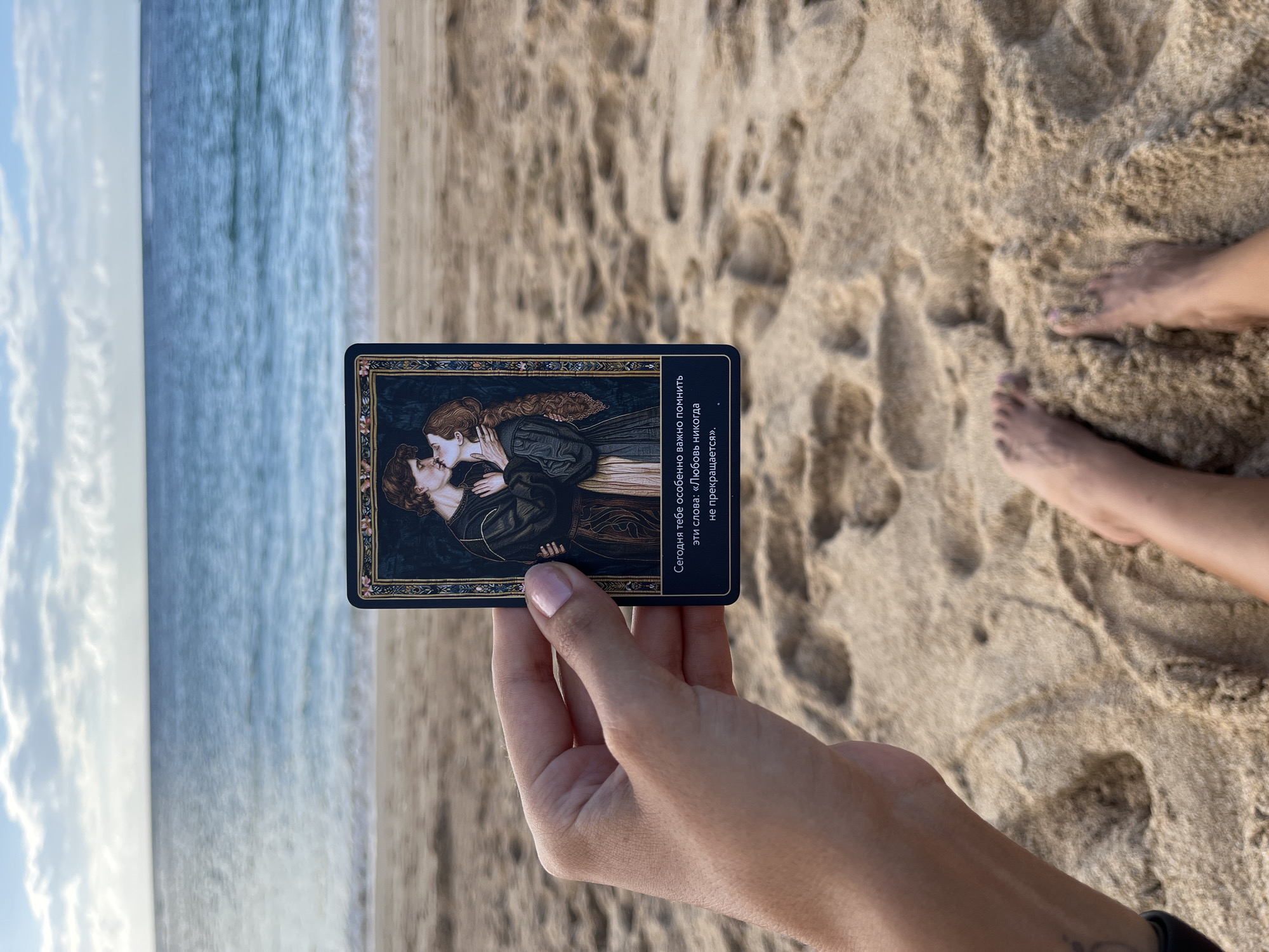 A person is holding a dark blue tarot card with an image of a couple kissing on a beach in Kuta Selatan, Indonesia. The card has gold trim with what appear to be flowers. The card has text in Russian, which translates to "Today it is especially important for you to remember these words: 'Love never ends.'" The hand holding the card has light skin and short, manicured fingernails. The background shows the beach sand and the ocean. There are two feet partially visible in the sand.