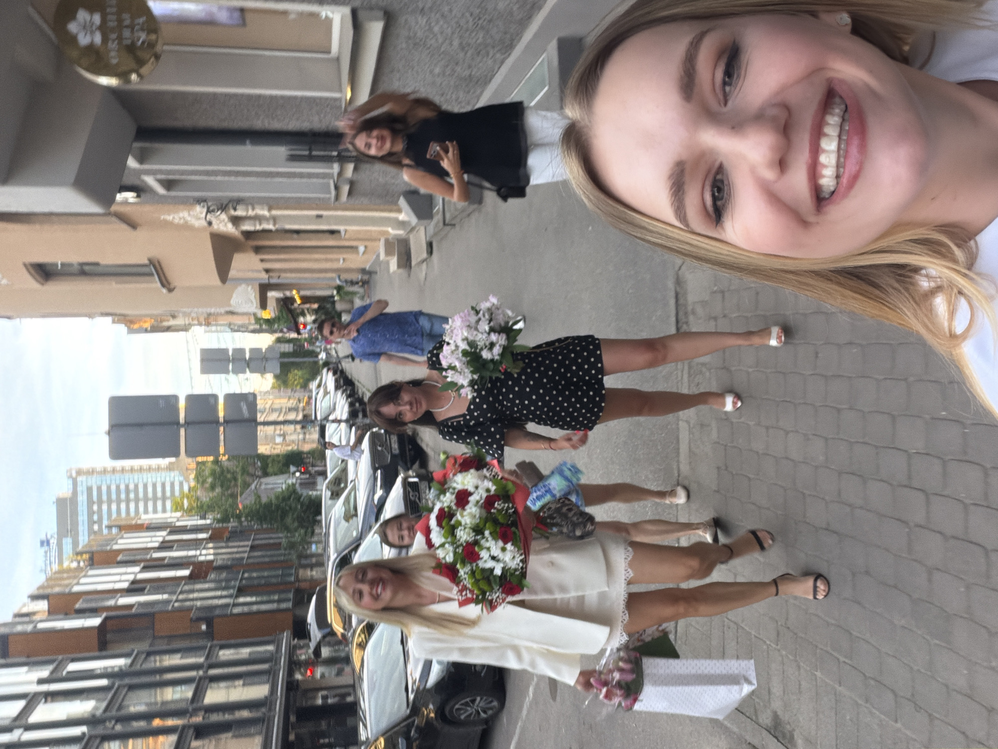 A group of four young women and one man are walking down a sidewalk in a European city. The woman in the foreground is taking a selfie, smiling broadly. She has blonde hair and is wearing makeup. The other three women are walking behind her. One is holding a bouquet of flowers and wearing a white blazer and skirt. Another is wearing a black polka dot dress and holding a bouquet of light pink flowers. The third woman is in the background, wearing a black top and white pants. The man is further back, wearing a blue shirt and jeans. They are walking on a sidewalk made of grey pavers, with buildings on either side of the street. Cars are parked along the street. The buildings are a mix of modern and older architecture. There are streetlights and signs visible. The sky is overcast. The setting appears to be a street in Riga, Latvia, given the presence of a sign for "Orchidea Thai SPA" which is a known business in Riga.