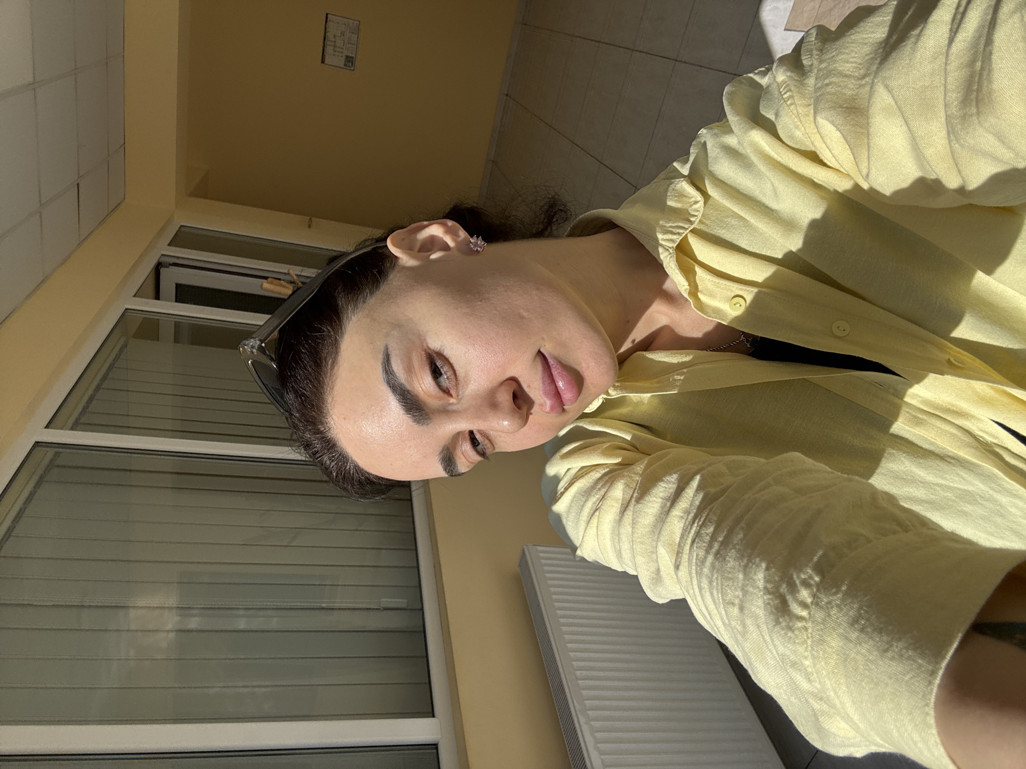 A medium-angle selfie shows a person indoors in a yellow shirt. In the background is a window with white trim and a white radiator. The location is unknown.