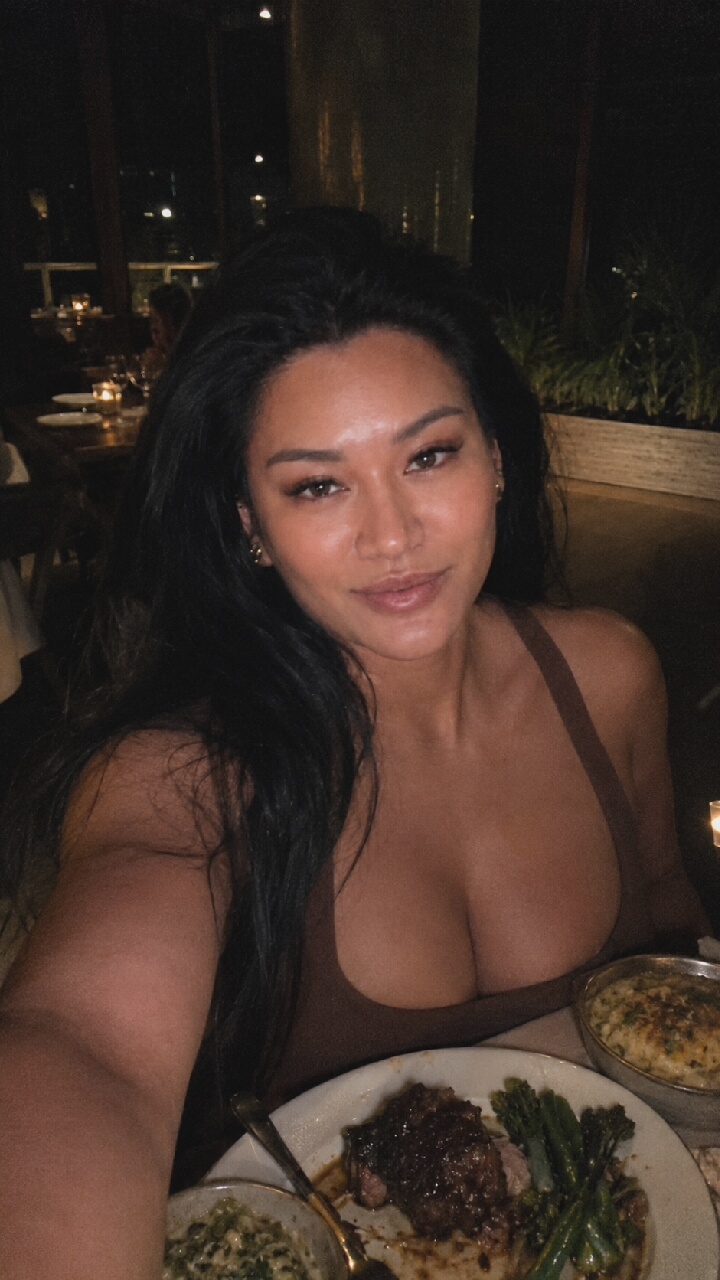 A woman with dark hair and light makeup is taking a selfie at a table in a restaurant. She is wearing a brown tank top and has her hair down. On the table in front of her is a plate of steak, asparagus, and a side dish, as well as another dish of what appears to be mac and cheese. The restaurant has a dimly lit, upscale ambiance with other patrons visible in the background. The image does not provide any information to suggest the location is Moscow, Russia.