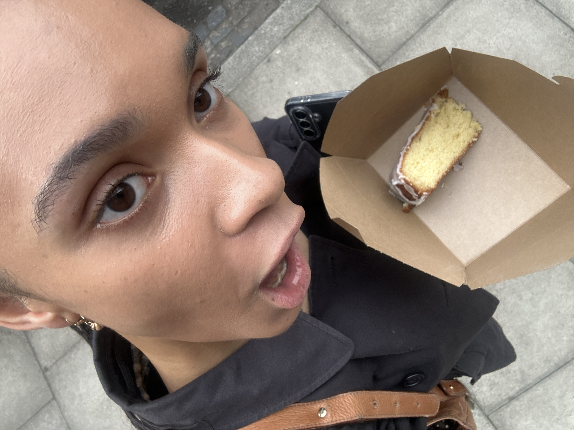 A close-up, high-angle shot shows a person holding a piece of cake in a cardboard container. The person, likely female based on facial features, has light brown skin, brown eyes, and thin eyebrows. She wears a black jacket and a brown leather belt. A phone is visible behind the cake. The location is suggested to be London, United Kingdom, based on the backdrop: grey paving stones. The subject seems to be capturing a selfie or photo of herself with the cake.