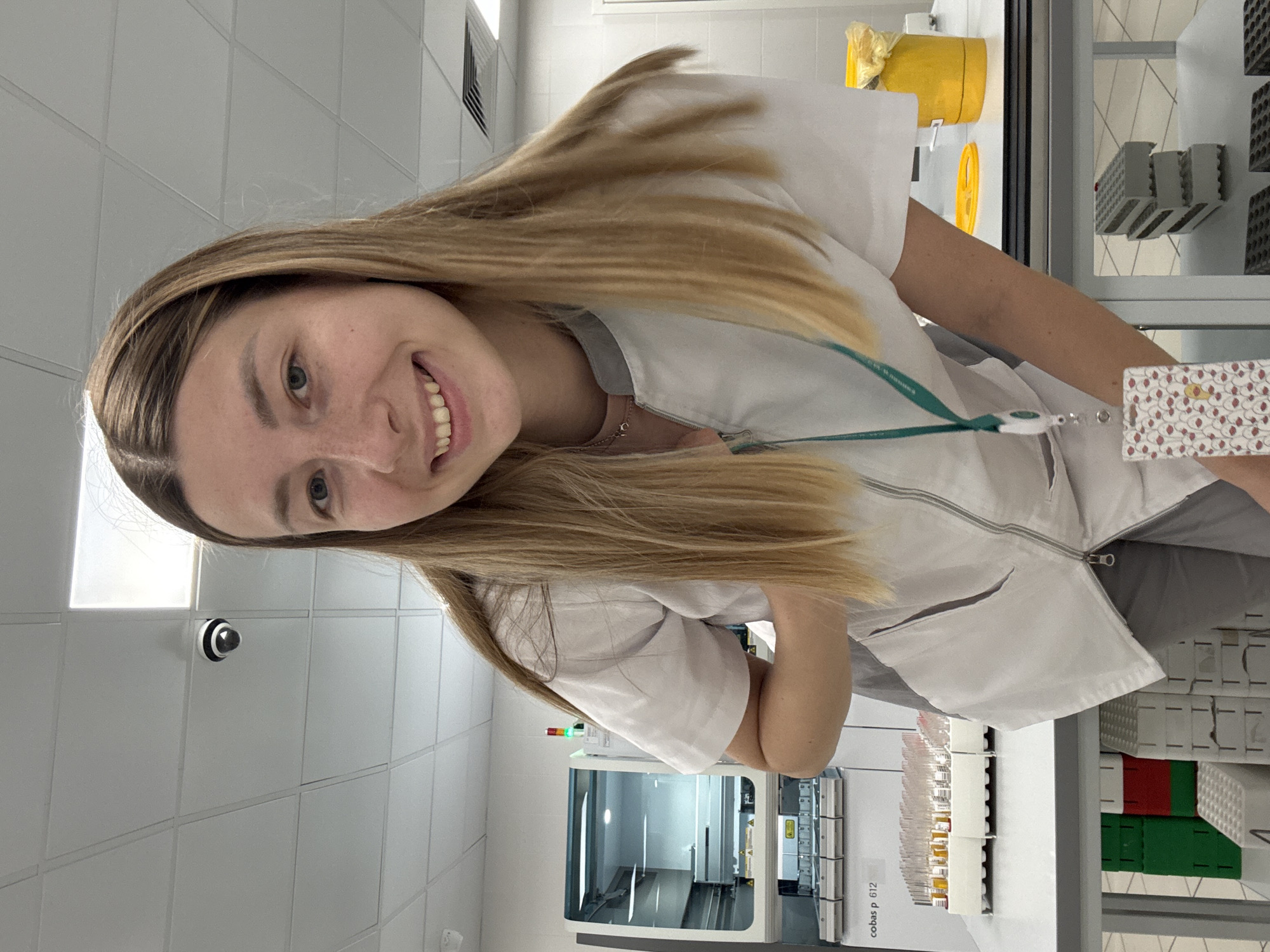 A young woman with blonde hair, wearing a white lab coat, smiles at the camera. She is in a laboratory setting, with various scientific equipment visible in the background, including racks of test tubes and a laboratory machine with a screen. A security camera is mounted on the tiled wall behind her. The overall scene suggests a professional or research environment. The lighting indicates it could be daytime, as the room is brightly lit. The woman appears happy and approachable. There is text on the lab machine which reads "cobas p 612".