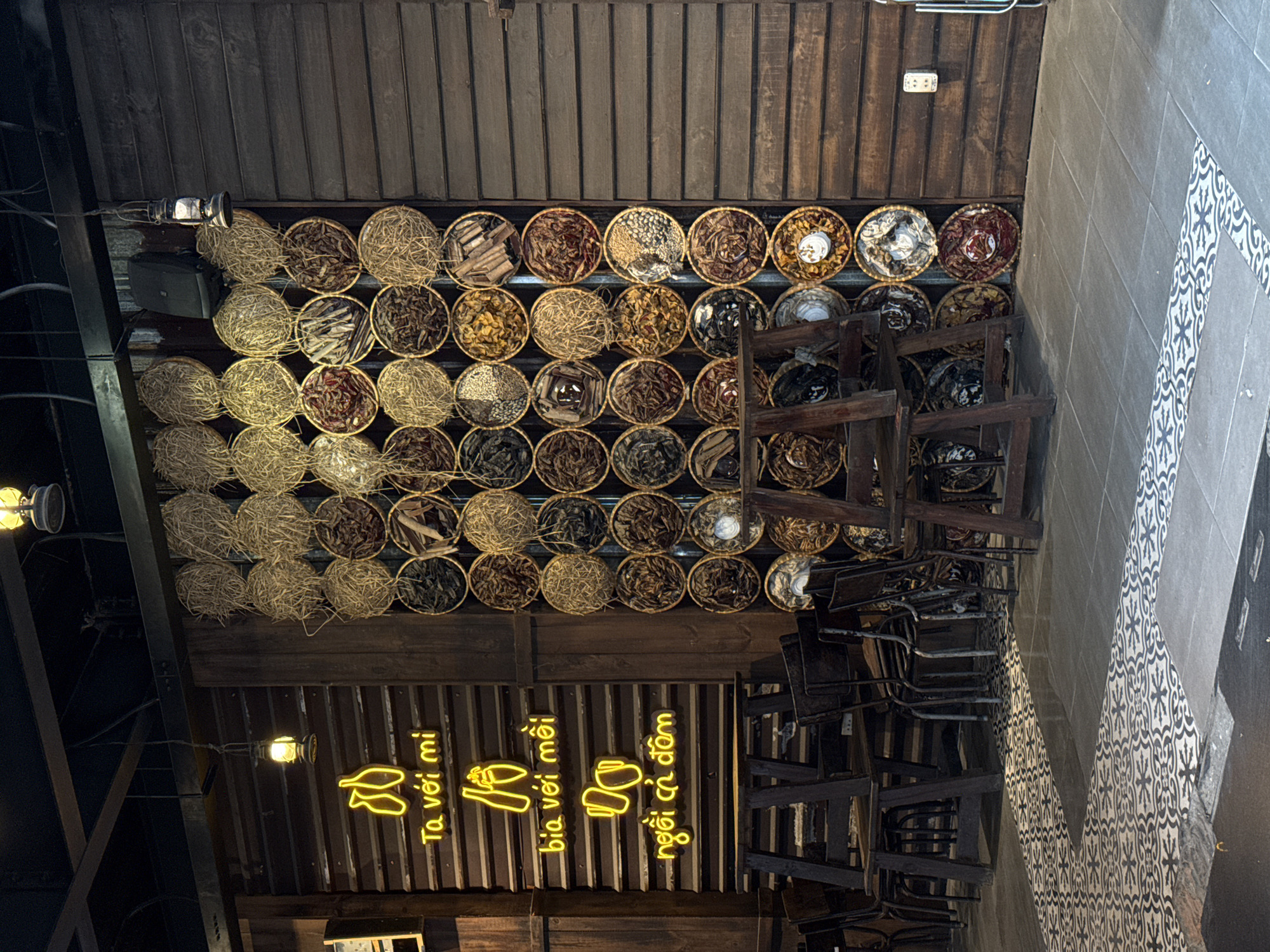 The image depicts a dimly lit interior space, likely a restaurant or bar, with a rustic and traditional Vietnamese aesthetic. The most prominent feature is a large wall adorned with numerous circular woven baskets, each filled with various dried goods such as herbs, spices, and grains. The baskets are arranged in a grid pattern, creating a visually striking display.
To the left of the display wall is a dark wooden wall with horizontal paneling, featuring yellow neon signs with Vietnamese text. The neon signs read "Ta với mì" (I with noodles), "bia với mồi" (beer with snacks), and "ngồi cà phê" (sit for coffee). Below these signs, a section of the wall appears to be corrugated metal.
In the foreground, there are several dark wooden chairs and tables, some of which are stacked or partially obscured. The floor is tiled with a distinctive pattern of black and white geometric and floral motifs.
The overall impression is one of a charming and authentic establishment, likely in a city like Đà Nẵng, Vietnam, where such traditional decorative elements are common. The lighting is atmospheric, with hanging lanterns and interior lights casting a warm glow on the scene. There are no people visible in the image.