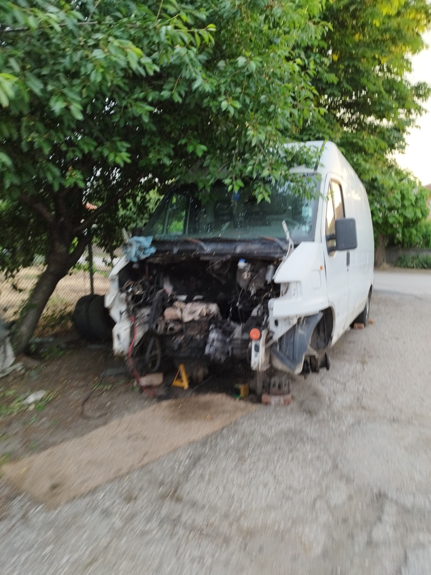 A white van is missing its front panels, tires, and engine parts and is sitting on concrete blocks and a yellow jack stand. There are two tires leaning against a tree next to the van. The van is parked on a gravel driveway under a tree.