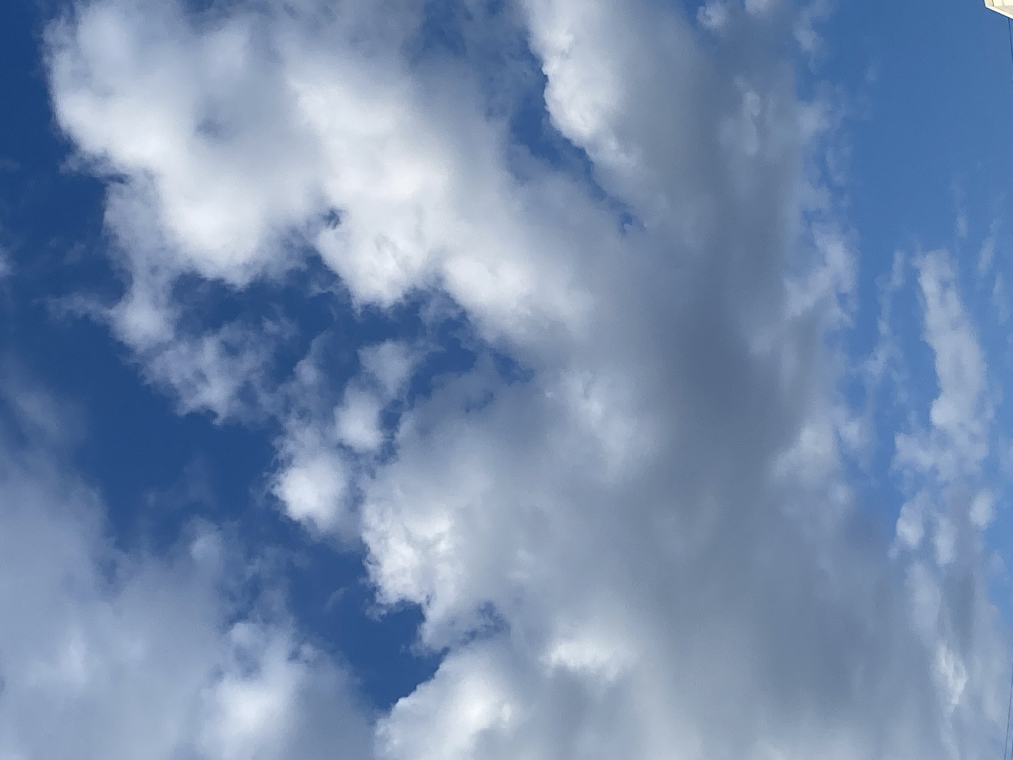 The image shows a sky filled with various clouds over the city of Molodizhne, Ukraine. The sky is a deep blue, with patches of white and gray clouds scattered across it. Some clouds appear fluffy and scattered, while others are larger and more dense, suggesting a mix of cumulus and possibly stratus clouds. The lighting suggests it is daytime, possibly late morning or afternoon, with the sun likely obscured by some of the denser cloud formations, casting shadows on other parts of the clouds. There are no people, subjects, or objects clearly visible in the frame, only the natural environment of the sky. The scene is tranquil and depicts a typical cloudy day.