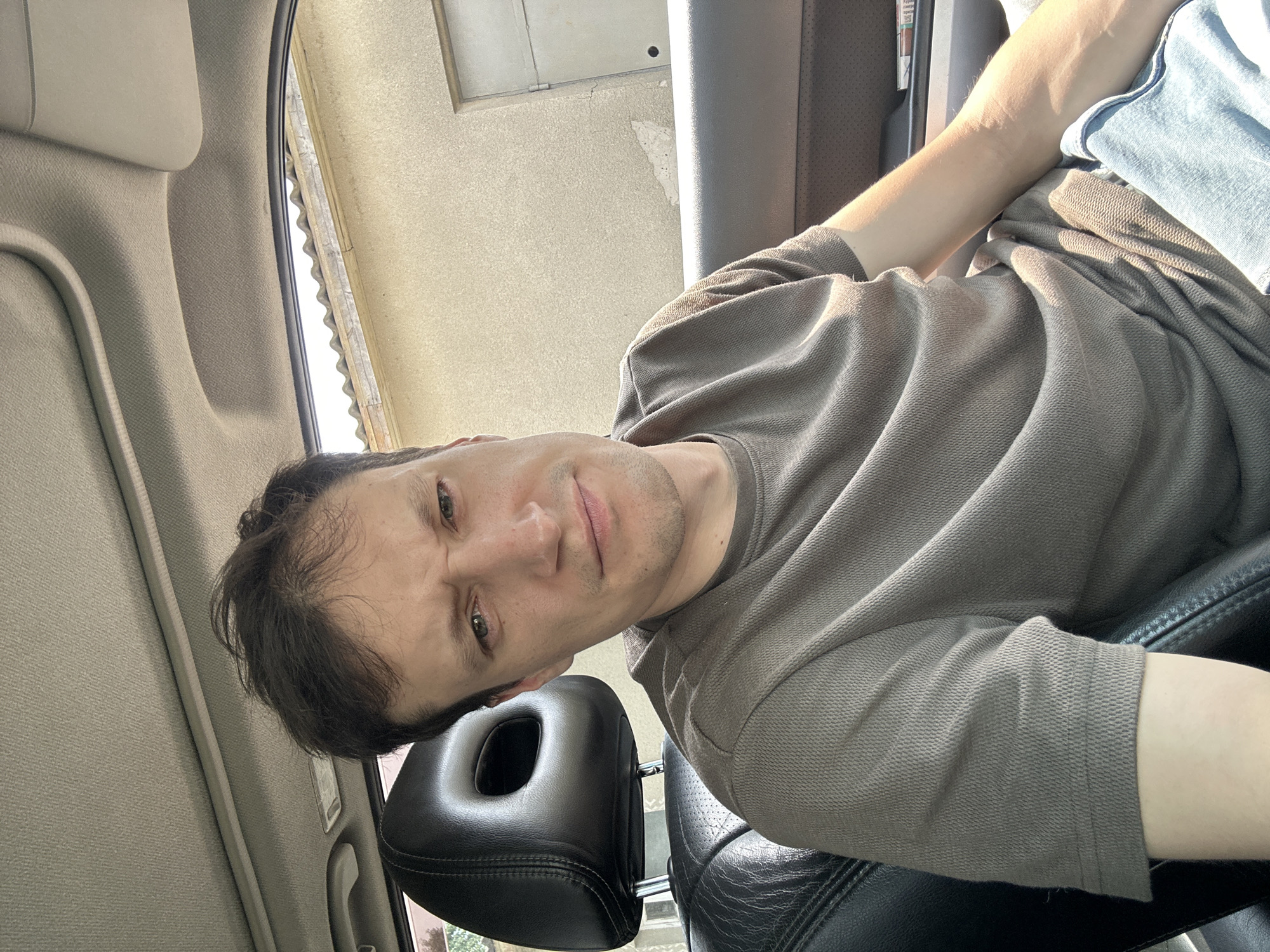 A man is seated in the driver's seat of a car, looking at the camera. He has dark hair and is wearing a gray t-shirt. The car's interior is visible, including the black leather seat and headrest. Outside the car, a light-colored building with a tiled roof is visible. The man's expression is a grimace, with furrowed brows. The scene is lit by natural sunlight.
