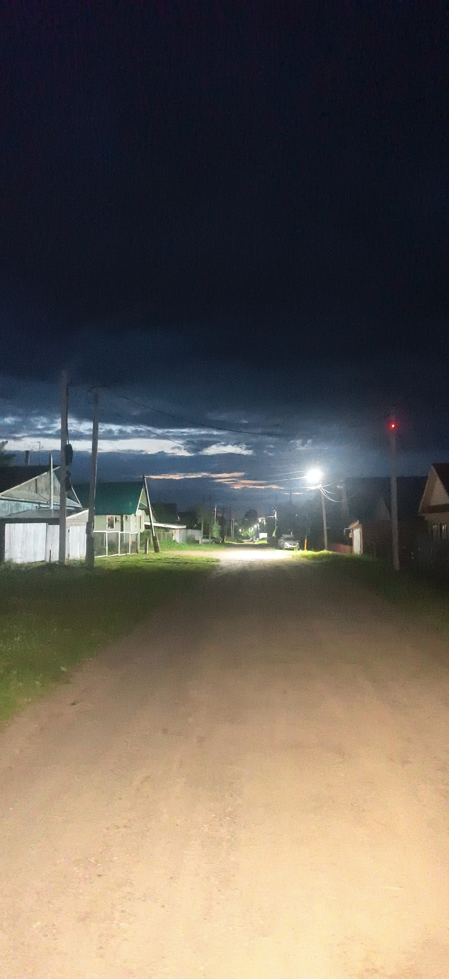 The image depicts a dirt road in the city of Fokino, Russia, at dusk or during twilight. The sky is dark with visible clouds, tinged with some light colors suggesting the last remnants of sunset. Along the sides of the road are small, rural-style houses with visible fences and grass. Several utility poles with wires are present, and one pole is illuminated by a streetlamp. Further down the road, the light from another streetlamp illuminates a parked car. There is a red light visible on a traffic signal or similar pole on the right side of the image. The road surface is rough and dusty, illuminated in the foreground by what appears to be headlights. No people are visible in the scene.