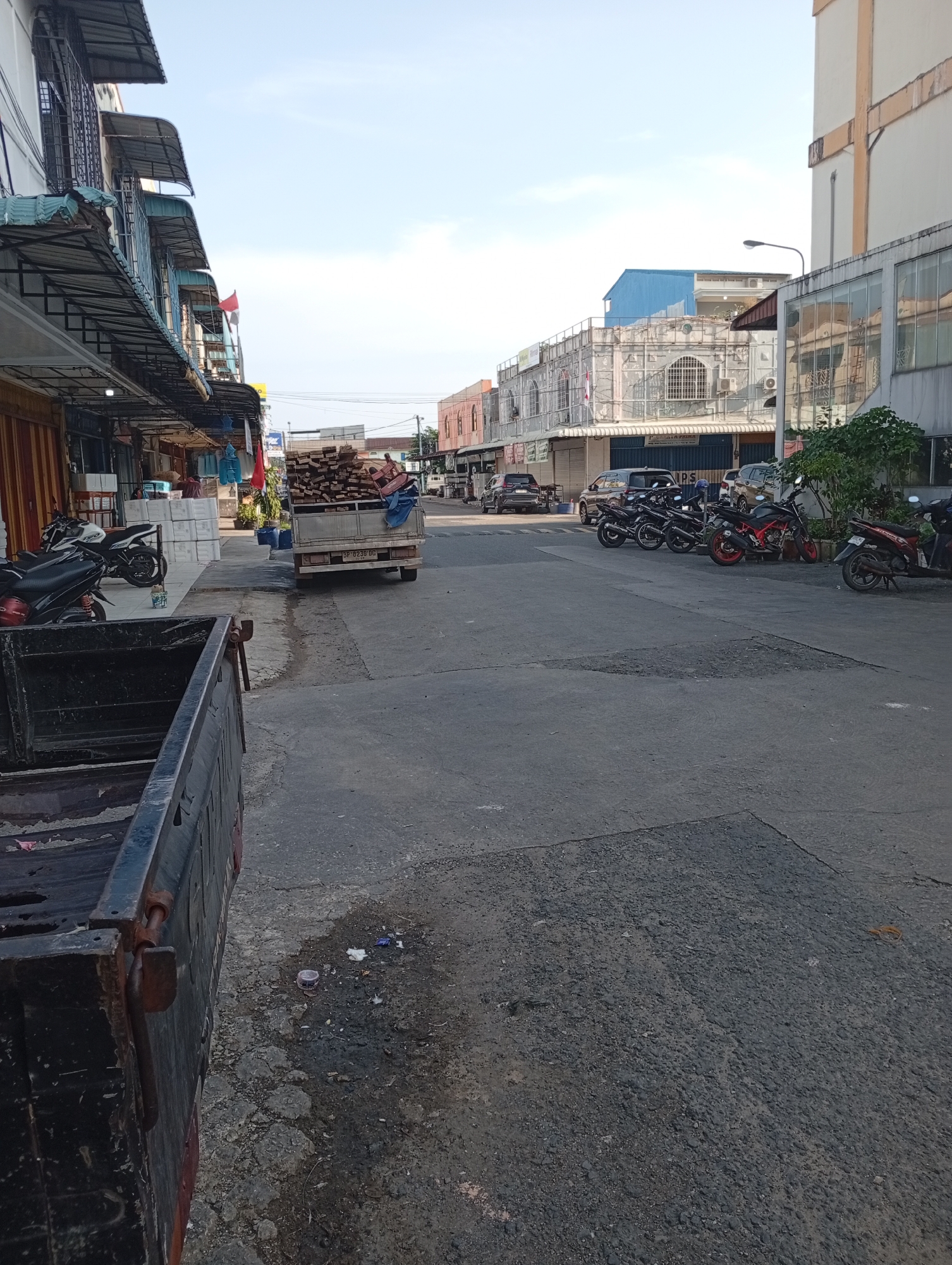 This image depicts a street scene in the Batam City, Indonesia area. On the left side of the frame, a row of single-story buildings with awnings lines the street. Several motorcycles are parked along the sidewalk in front of these buildings. In the center of the image, a flatbed truck is stopped, loaded with what appears to be wood. Further down the street, more motorcycles are parked along the right side. The road surface is asphalt, showing signs of wear and tear. The sky is clear with some clouds, suggesting it is daytime. The overall impression is of a commercial or residential street with active local transport.
