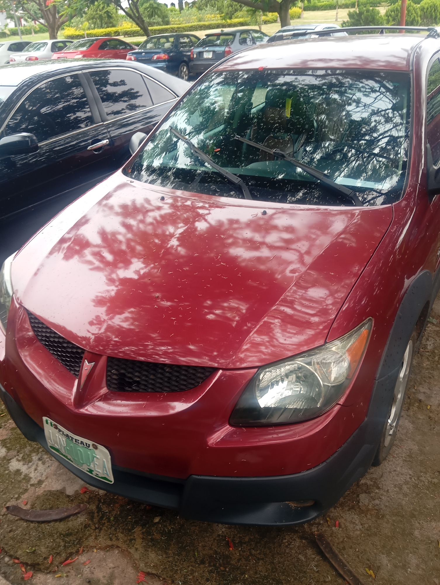 A red 2003-2008 Pontiac Vibe station wagon is parked on a wet, muddy, and leaf-strewn paved area, likely in the city of Jos, Nigeria, indicated by the "PLATEAU" inscription on its license plate, which reads "JJN-104EA." The car's hood is speckled with water droplets and reflections of trees. The front of the car is visible, including the grille, headlights, and bumper. A dark-colored sedan is partially visible to the left of the Vibe, and other vehicles are parked further back. The environment suggests an outdoor parking lot or roadside location with overgrown vegetation and trees in the background.