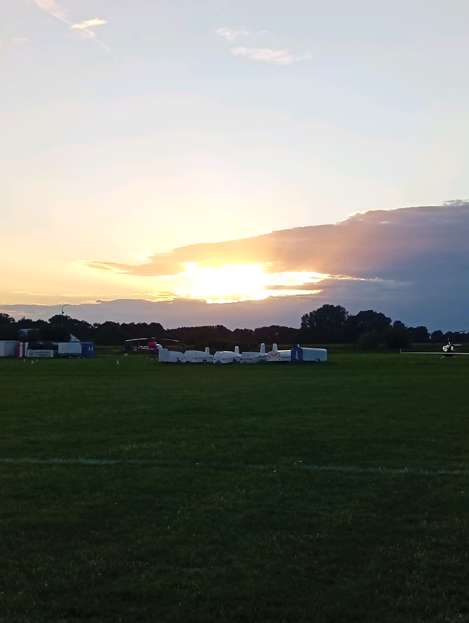 The image depicts a sunset over an airfield in Szewce, Poland. The sky is a mix of light blue and orange hues, with clouds partially obscuring the setting sun. In the foreground, a vast expanse of green grass covers the airfield, with a white line marking a boundary or path.
Several small aircraft, possibly gliders or ultralights, are parked on the tarmac in the middle ground. Some appear covered, while others are uncovered, revealing their sleek, aerodynamic shapes. To the left, there are a few small buildings or trailers, one red and white, and some orange traffic cones. A lamppost stands near these structures.
In the background, a dense line of dark green trees forms the horizon. Further in the distance, a red helicopter is visible. The overall scene is tranquil, suggesting the end of a day of aviation activities. The lighting indicates it is either early morning or late evening. There are no people visible in the frame.