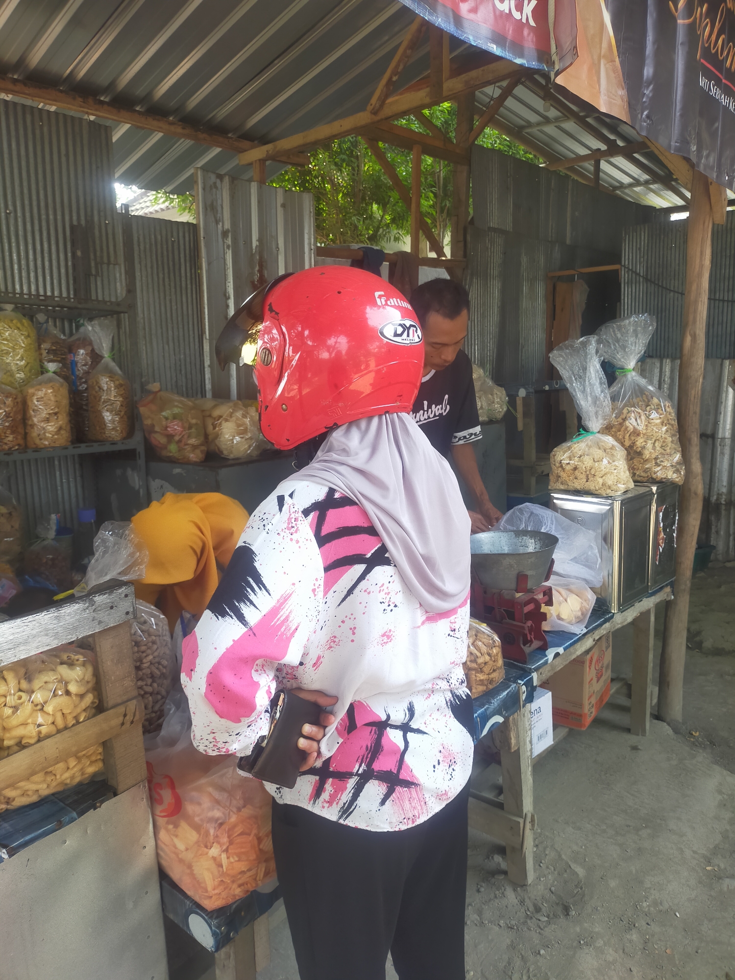 In Kalisapu, Indonesia, a person wearing a red helmet and a light purple hijab is standing with their back to the camera in front of a stall. They are wearing a white long-sleeved shirt with a pink and black abstract design and black pants. The person is holding a brown wallet in their right hand. Behind them, a man in a black t-shirt is visible, facing down towards a counter. The stall is constructed with corrugated metal and wood. Shelves behind the man are stocked with various bagged food items. On the counter in front of the man, a vintage red and silver scale is present, along with bags of food and what appear to be metal canisters. The ground is a rough, gray concrete surface.
