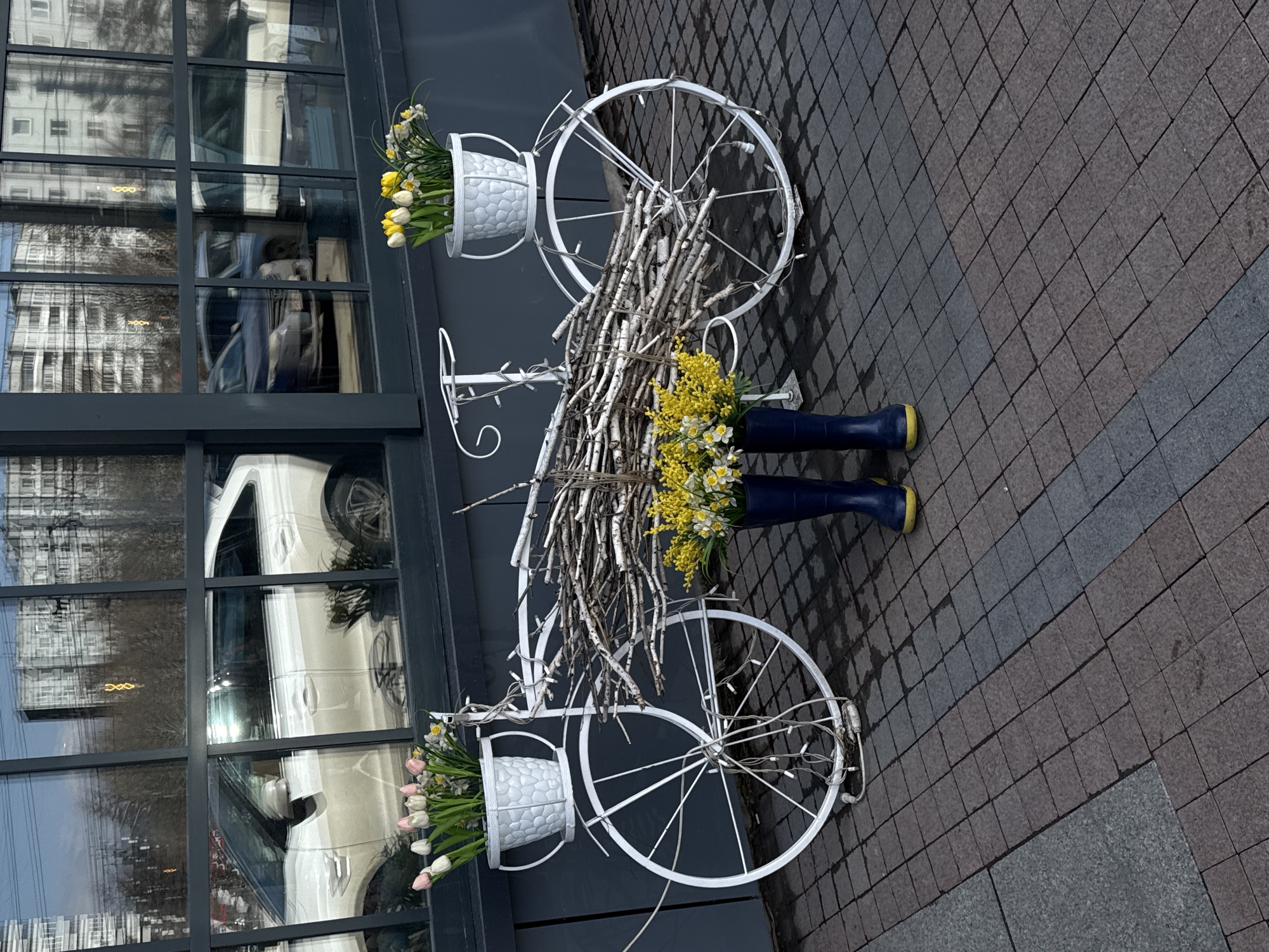 A decorative white bicycle with flower baskets stands in front of a building in Krasnoyarsk, Russia. The bicycle has baskets on the front and back, each filled with flowers. The handlebars are wrapped in branches. Blue rain boots with yellow soles are placed near the center of the bicycle, also holding flowers. The building behind the bicycle has large windows that reflect a building and a white car. The bicycle sits on a tiled sidewalk.