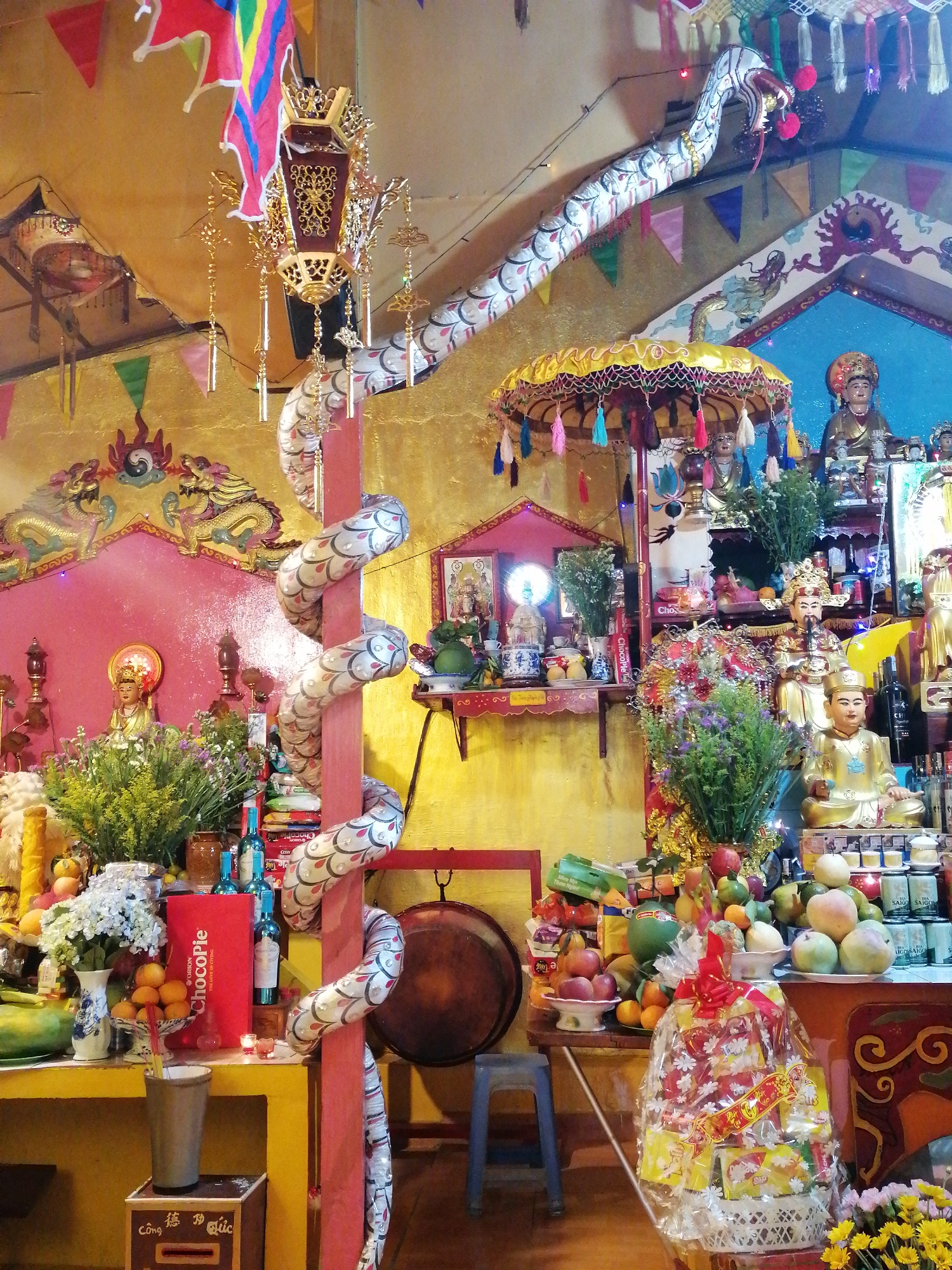 This vibrant image captures the interior of a richly decorated temple or shrine, likely in Vietnam. The walls are painted in striking gold and pink/red hues, adorned with intricate dragon and cloud patterns, while colorful bunting flags hang from the ceiling.
The scene is dominated by multiple altars laden with numerous offerings and statues. Golden figures of deities or revered individuals, some resembling Buddhist Bodhisattvas, are prominently displayed. A distinctive, long, white serpent or dragon figure, patterned with scales, wraps around a red wooden pillar and extends towards the ceiling, its head featuring a red snout. An ornate golden lantern with delicate hanging charms is suspended from above, and a yellow, fringed umbrella with colorful tassels shades one of the altars.
Offerings include copious amounts of fresh fruits such as mangoes, apples, oranges, and papayas, carefully arranged in bowls and platters. Fresh floral arrangements of purple, white, and yellow flowers, alongside green foliage, add to the vibrant display. Packaged items like bottles of drinks (some blue, some clear glass), cans of "Saigon Special" beer, and a large red box labeled "ChocoPie" are also present. A large cellophane-wrapped gift basket, filled with yellow-packaged snacks, sits on the floor. An incense holder with burning sticks suggests ongoing devotional practices.
Other details include a large, round bronze gong hanging on the right, and a wooden box on the floor inscribed with "Công Đức," indicating a donation box for merit or good deeds. The overall atmosphere conveys a strong sense of reverence and cultural tradition, characteristic of a Vietnamese place of worship. The blend of traditional religious iconography with modern packaged goods reflects the evolving nature of offerings.