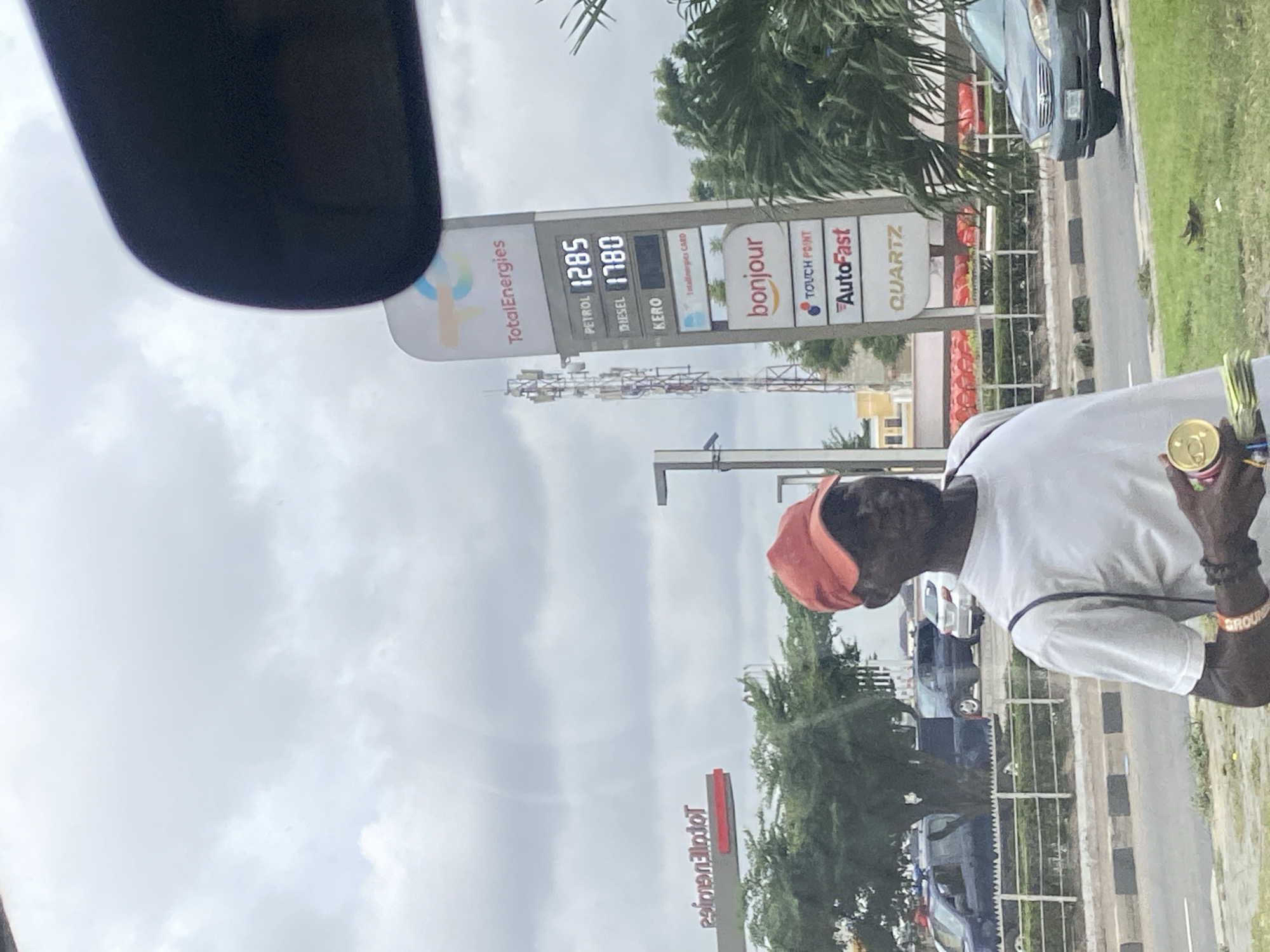 In Ikeja, Nigeria, a man wearing a white t-shirt and an orange cap stands on the side of a road, holding a can and some folded bills. Behind him, a TotalEnergies gas station sign displays fuel prices, with "Petrol" at 1285 and "Diesel" at 1780. Further back, a cell tower and several parked vehicles are visible, including a dark blue police vehicle. The sky is overcast, suggesting cloudy weather.