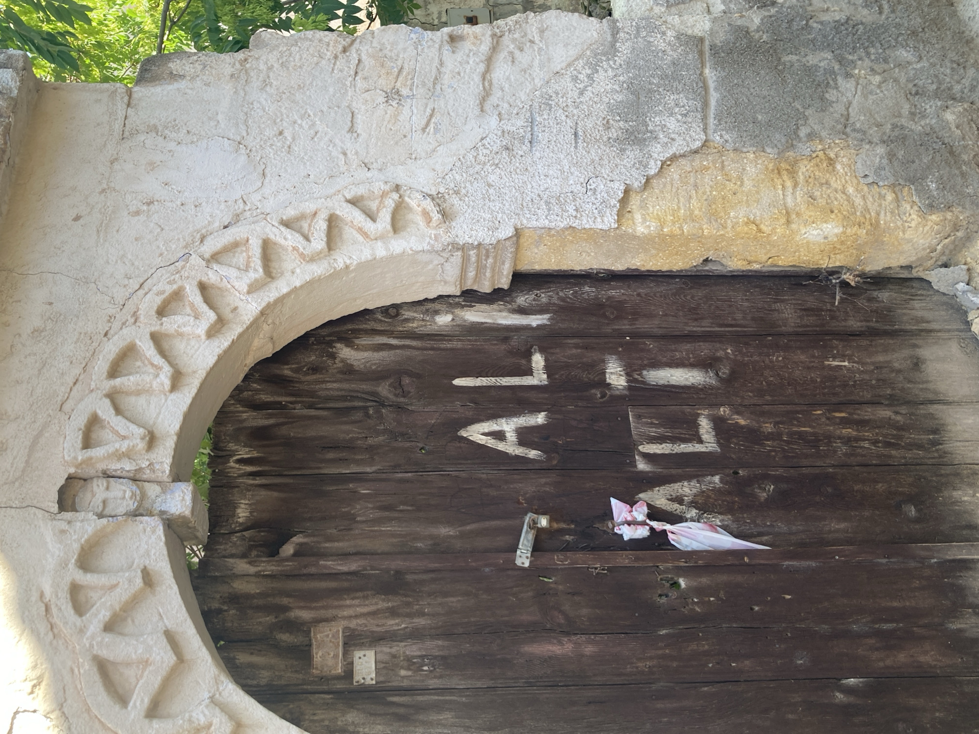 In the city of Avanos, Turkey, an old wooden door is set within a weathered stone archway. The arch is adorned with intricate carvings, including a small, stylized face. The door itself is dark brown and shows signs of age and wear. A metal latch is visible, secured with a pink and white ribbon. White graffiti, spelling out "AL" and "ALT," is scrawled across the door's surface. The surrounding stone wall is crumbling in places, revealing older construction beneath. Lush green foliage is visible in the background, suggesting an outdoor setting.