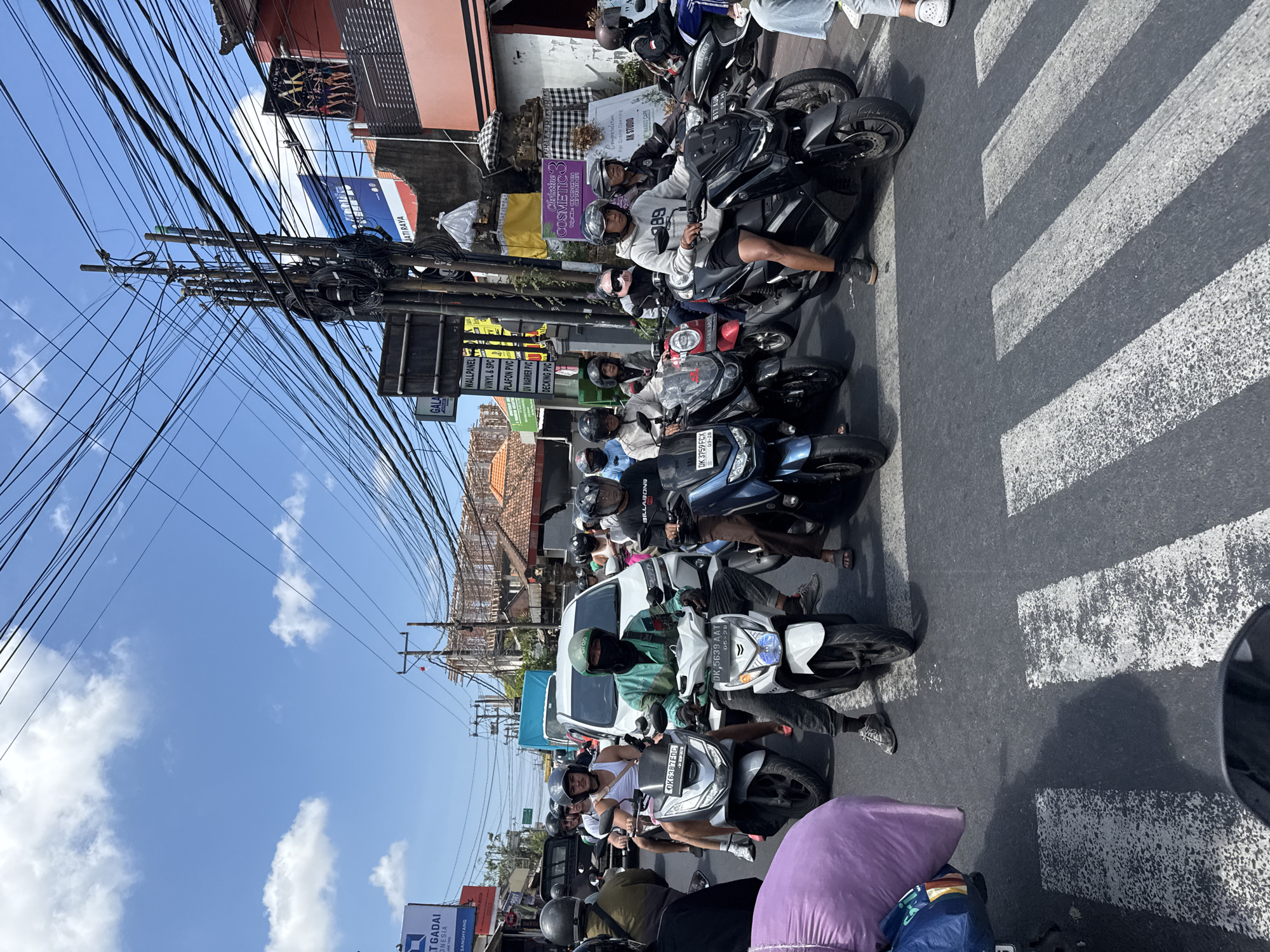 The image captures a busy street scene in North Kuta, Indonesia, with a significant amount of motorcycle traffic. Numerous individuals on motorcycles, many wearing helmets, are navigating the road. Some are seen riding alone, while others have passengers. The environment is characterized by a clear blue sky with scattered clouds, and a dense network of overhead electrical wires. Buildings line the street, some featuring signage for businesses. The road itself is paved, with a pedestrian crossing marked by white stripes. The overall impression is one of a bustling urban environment with a high volume of motorcycle transportation.