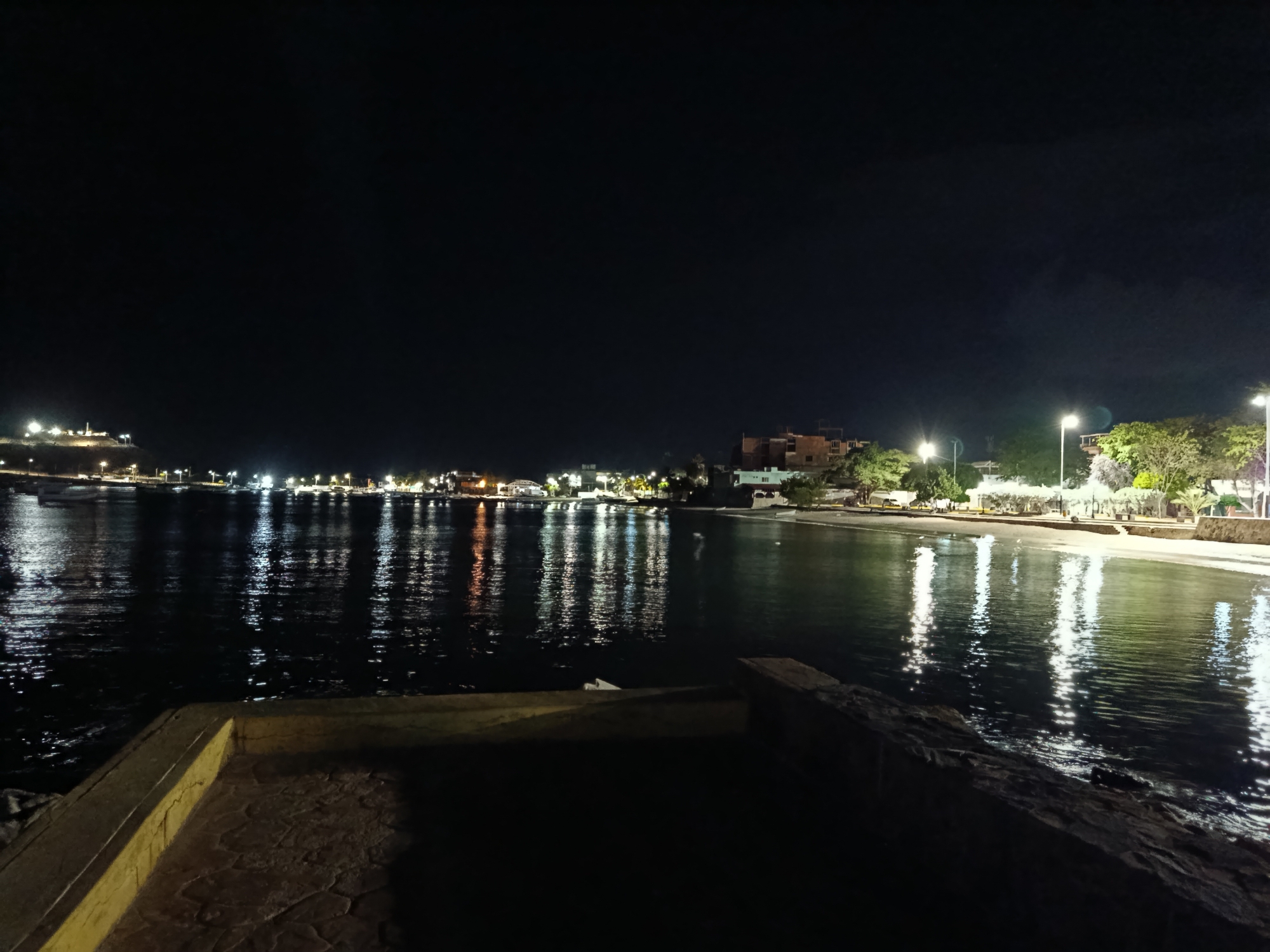 The image depicts a nighttime scene of a coastal area, likely in Juan Griego, Venezuela, characterized by the dark sky and illuminated waterfront. The foreground features a paved walkway and a low stone wall bordering the water. The water itself reflects the lights from the buildings and streetlights along the shoreline, creating a shimmering effect. In the mid-ground, a town or village is visible with numerous lights indicating buildings and streetlamps. A beach area can be discerned, with trees and vegetation lining the shore. Several boats are present in the water, their lights also contributing to the reflections. The overall atmosphere is tranquil and illuminated, suggesting a populated coastal settlement at night.