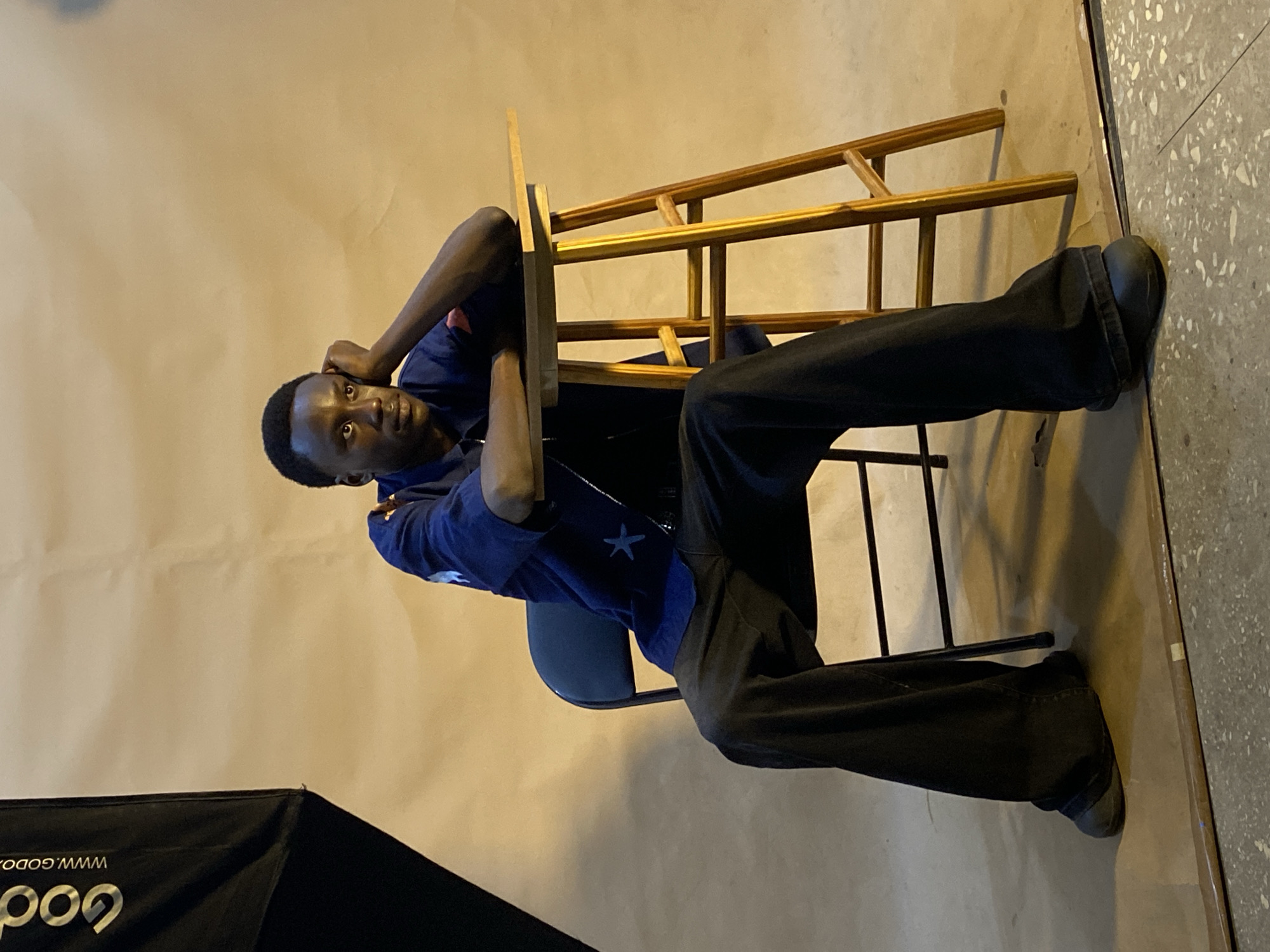 In Gura Top, Nigeria, a Black man sits on a black folding chair next to a small wooden stool. He is wearing a dark blue shirt with star patterns and dark wide-leg pants. His right arm rests on the stool's surface, and his head is propped on his hand, looking off to the side with a pensive expression. The background is a plain, tan-colored studio backdrop, with a large softbox light modifier visible on the left side of the frame. The floor appears to be concrete with some debris.