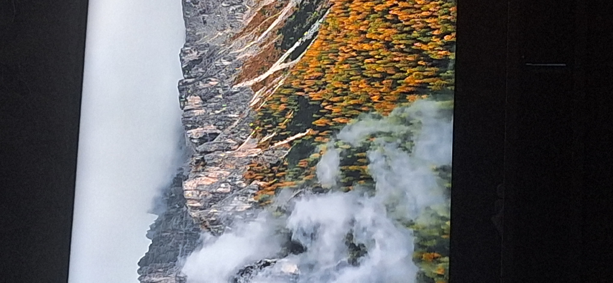 The image shows a mountainous landscape with trees in autumn colors and fog. The mountains are rugged and rocky, with some patches of snow or ice. The trees are densely packed on the lower slopes and are a mix of green, yellow, and orange, indicating the fall season. Wisps of fog are drifting through the scene, obscuring some of the details of the mountains and the forest. The sky is overcast and gray. The image is taken from a distance, giving a wide view of the natural scenery. There is no indication of the city of Bila Tserkva, Ukraine, in this image.