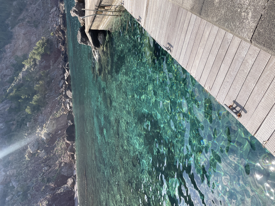 The image depicts a coastal scene at a cove in Valldemossa, Spain. A clear, turquoise-green body of water occupies the left and central portions of the frame, revealing the rocky seabed through its transparent surface, with sunlight reflections shimmering across the ripples. Along the right side, a light-colored wooden dock with horizontal ridged planks extends from the foreground into the midground. Two grey metal boat cleats are affixed to this wooden surface. To the right of the wooden dock, a darker concrete or stone surface forms part of the pier structure. In the background, where the dock meets the shore, a metal ladder with multiple rungs descends into the water from a stone embankment. The background features steep, rugged cliffs composed of reddish-brown rock, with sparse green vegetation, including pine trees, growing on their slopes. Large dark boulders are visible both on the shoreline and partially submerged in the water along the base of the cliffs. A distinct sunbeam is visible in the upper left, casting light onto the cliff face. No individuals, animals, or specific actions are depicted in the scene.