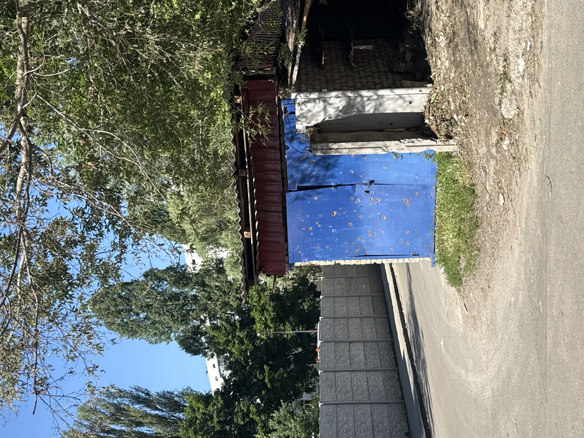 A bright blue garage door, riddled with shrapnel marks, dominates the foreground of the image. To the right of the door, a partially constructed brick wall is visible, showing signs of damage and incomplete construction. The garage is situated next to a paved road that curves to the left. Behind the garage, a grey concrete fence stretches into the distance. The scene is framed by lush green trees, with their leaves casting dappled shadows across the ground. In the background, several tall buildings can be seen peeking through the foliage, suggesting an urban or suburban setting. The sky is clear and blue, indicating a sunny day. The overall impression is one of a quiet, somewhat neglected area, with the shrapnel marks on the garage door hinting at a past conflict. The location is identified as Kyiv, Ukraine.