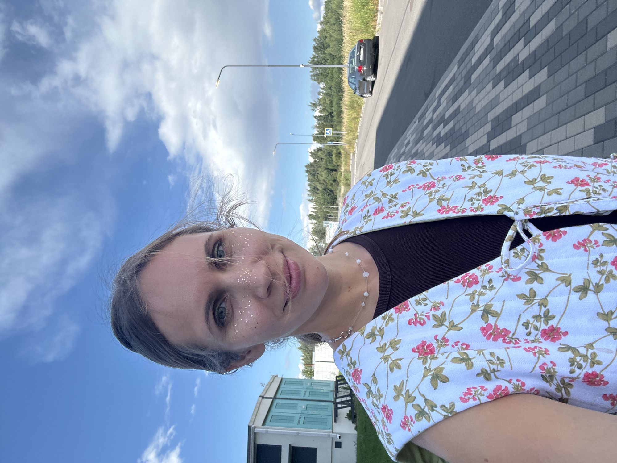 A woman with blonde hair tied back is taking a selfie outdoors. She is wearing a white vest with a pink floral pattern over a dark brown top, and a delicate necklace with small beads. She has glitter applied to her cheeks. The background shows a modern building on the left, a paved walkway with gray and black bricks, a road with a dark-colored car parked on the right, streetlights, and a line of trees and tall grass. The sky is blue with white clouds. The location is likely an industrial or commercial area in Vsevolozhsk, Russia.