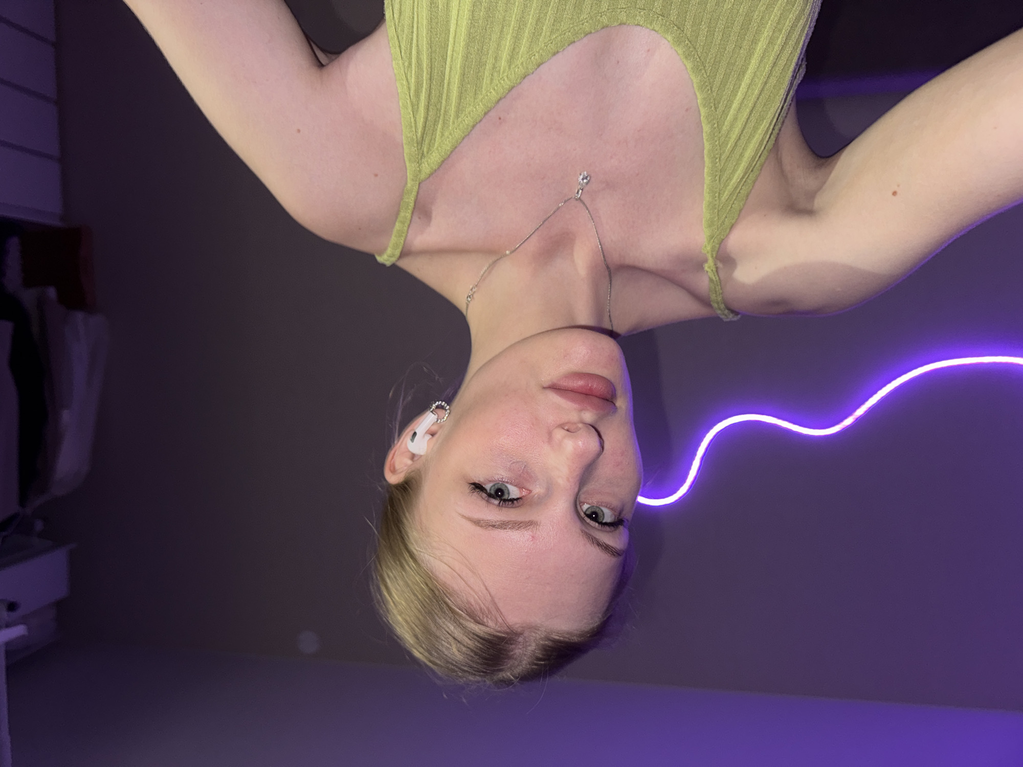 A young Caucasian woman with blonde hair pulled back and light blue eyes is looking directly at the camera. She is wearing a light green ribbed camisole and a silver pendant necklace with a single clear stone. She has white wireless earbuds in her right ear. The background is a purple wall with a neon purple light strip running horizontally across it. To the right of the woman, a closet or wardrobe is visible with some clothing hanging inside and a white drawer unit. The lighting suggests an indoor, dimly lit environment, possibly a bedroom or dressing area. There is no indication of the city of Perm, Russia, or any specific urban location.