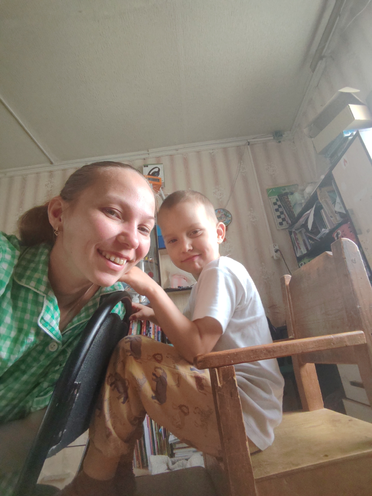 A woman and a young boy are indoors in Чухлэм, Russia. The woman is holding a black chair and smiling at the camera, wearing a green and white checkered top. The boy is seated on a small wooden chair, wearing a white t-shirt and patterned pajamas. Behind them is a shelf with books and other items. The walls are decorated with wallpaper, and there is a window on the right side of the image. The ceiling is white.