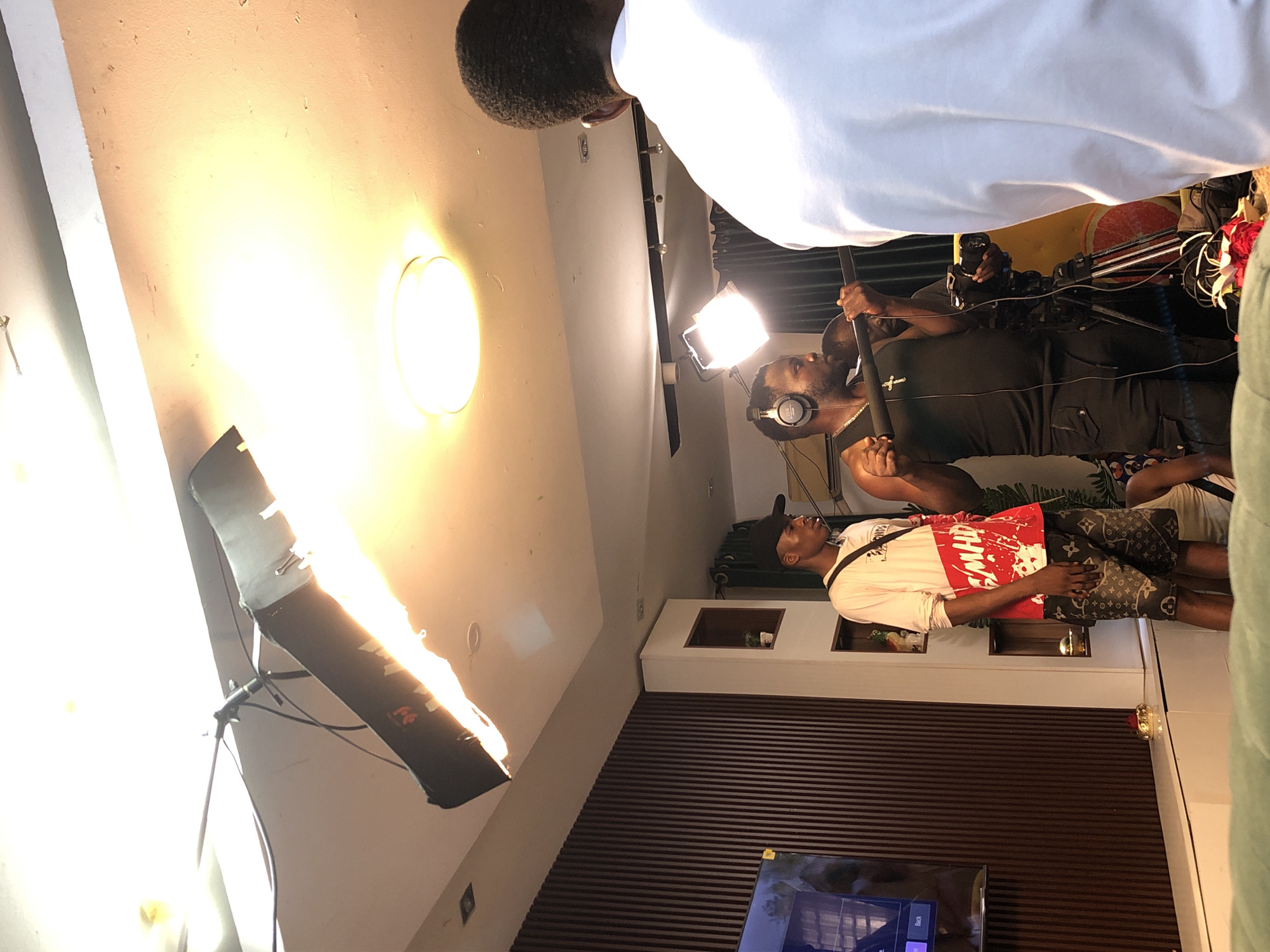 The image captures a scene in what appears to be an indoor setting, potentially a studio or a room being used for filming. A person wearing a white shirt, a red and white patterned upper garment, and patterned shorts is visible. He is standing near a wall with dark horizontal stripes and a decorative element on it.
A person with headphones is operating what looks like a camera or a microphone boom, and there is a video camera visible in the background. Other lighting equipment, including a softbox light on a stand, is present, indicating the setup for a video shoot.
The location is listed as being in the city of Unknown, Unknown.