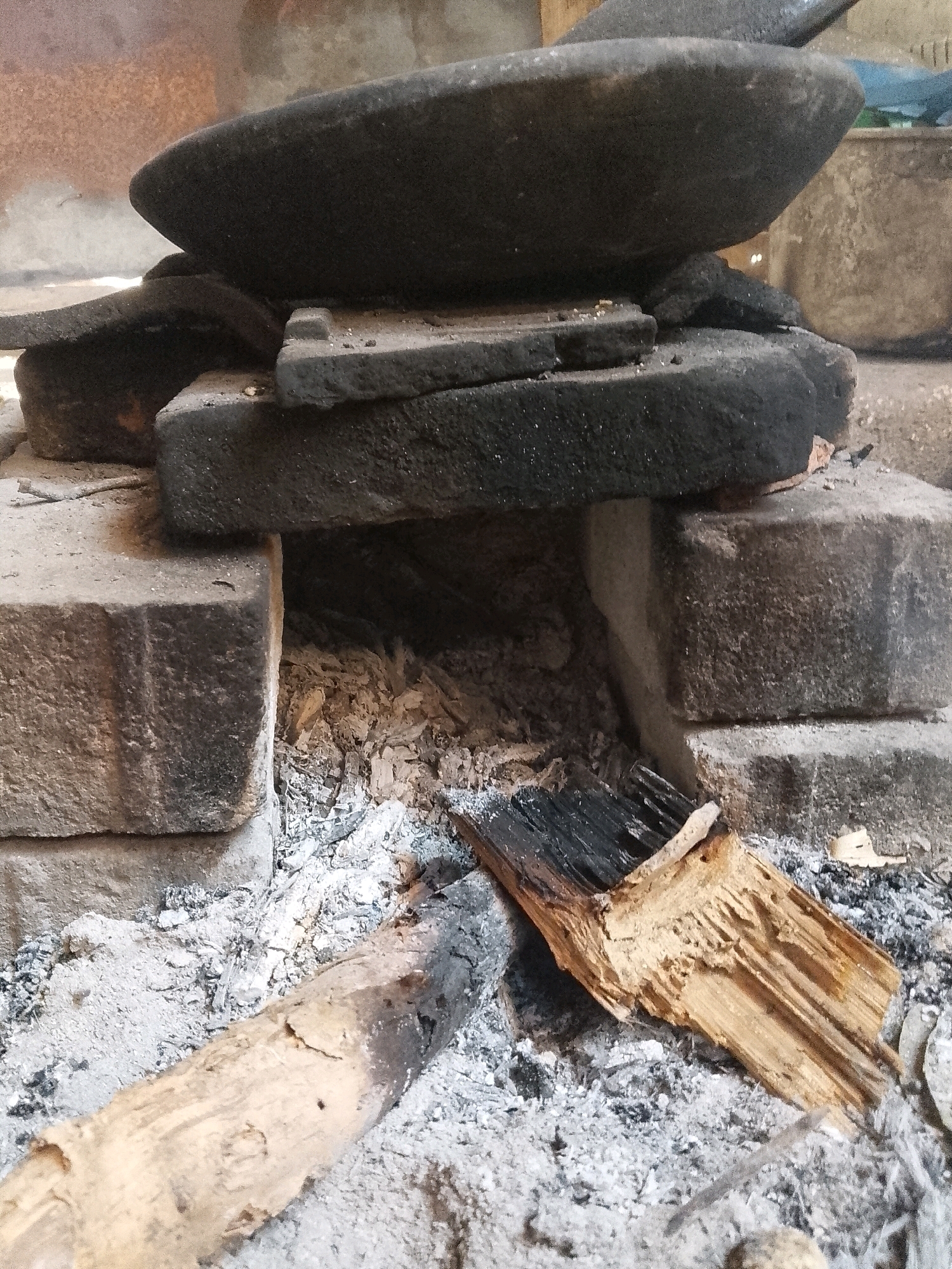 The image depicts a close-up view of a traditional, rustic hearth or stove likely found in Wadang, Indonesia. The primary focus is on the construction of the stove, which appears to be made from rough, rectangular bricks. On top of these bricks rests a large, dark, concave stone, possibly a grinding stone or a cooking surface, supported by smaller, flat, dark stones. Below this structure, a bed of ash and embers is visible, indicating recent or ongoing use. Two pieces of wood are present in the foreground, one a long, light-colored log partially covered in ash, and the other a more charred and broken piece of wood, showing signs of combustion. The overall impression is one of a functional, utilitarian cooking setup, likely situated outdoors or in a simple, open structure, given the surrounding materials and the presence of raw firewood. The context suggests a traditional Indonesian setting where such methods of cooking are still in practice.