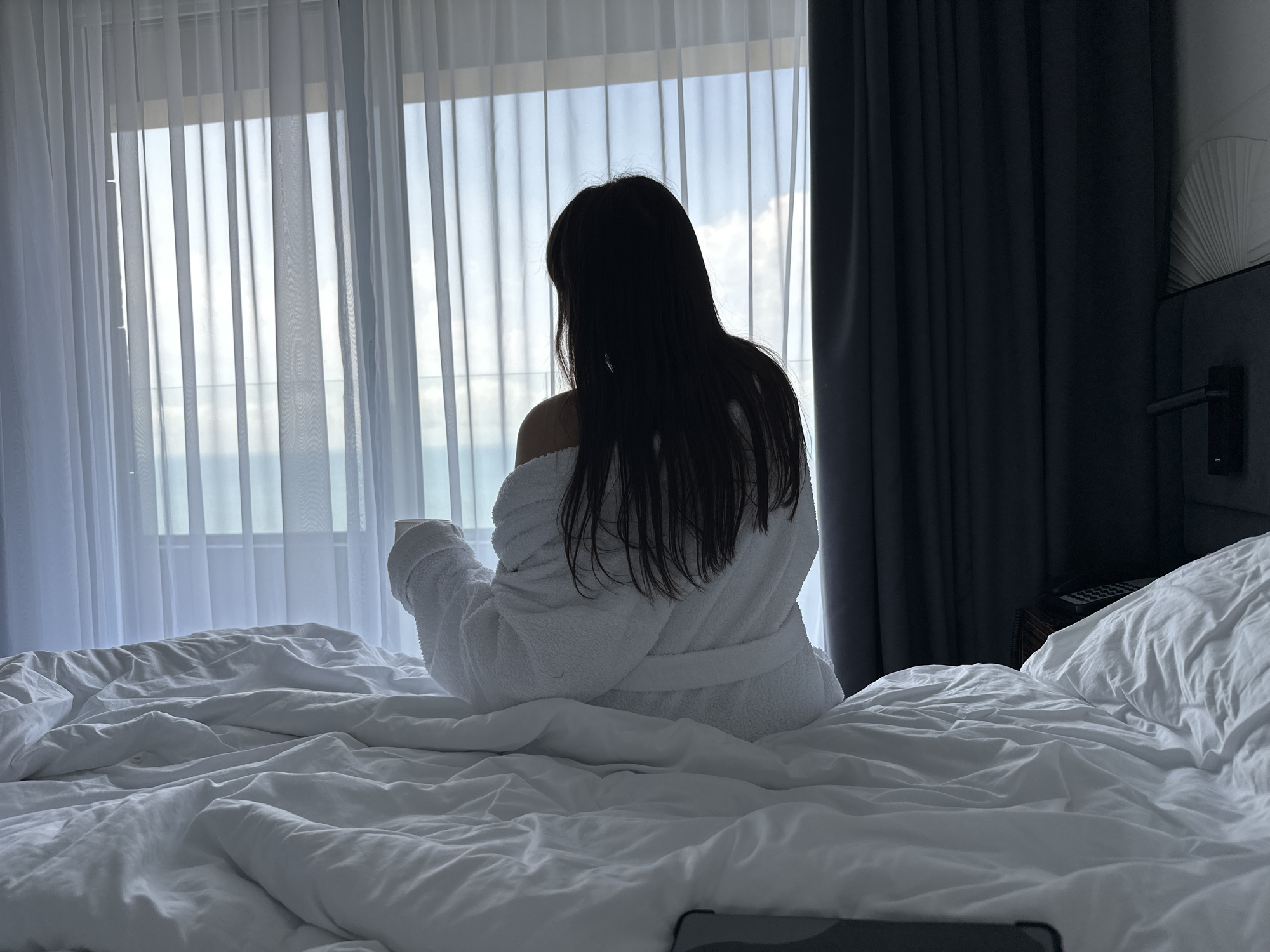 A woman is sitting on a bed, facing a window. She is wearing a white bathrobe. The bed is covered in white sheets and a white comforter. There is a window behind the woman. The window is covered with thin, white curtains. Through the window, a view of a building and the horizon can be seen. To the right of the window are thick, dark grey curtains. The edge of the headboard is visible. The scene suggests the interior of a room in a hotel in Sochi, Russia.